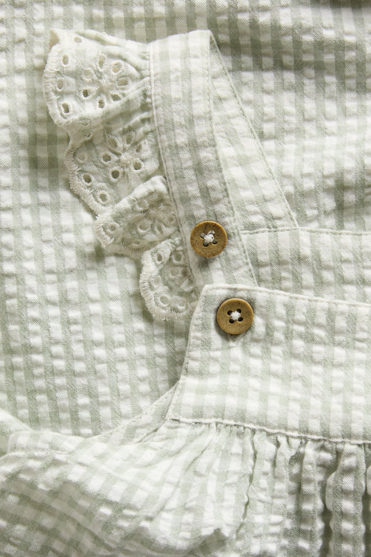 Zbliżenie na zieloną, kraciastą sukienkę niemowlęcą z haftowanymi falbankami i drewnianymi guzikami.
