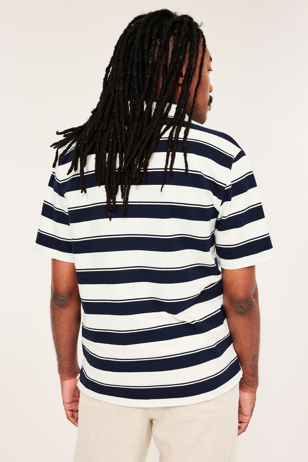 Mies takaa, yllään tummansinivalkoraidallinen lyhythihainen pikeepaita ja vaaleanbeiget housut.