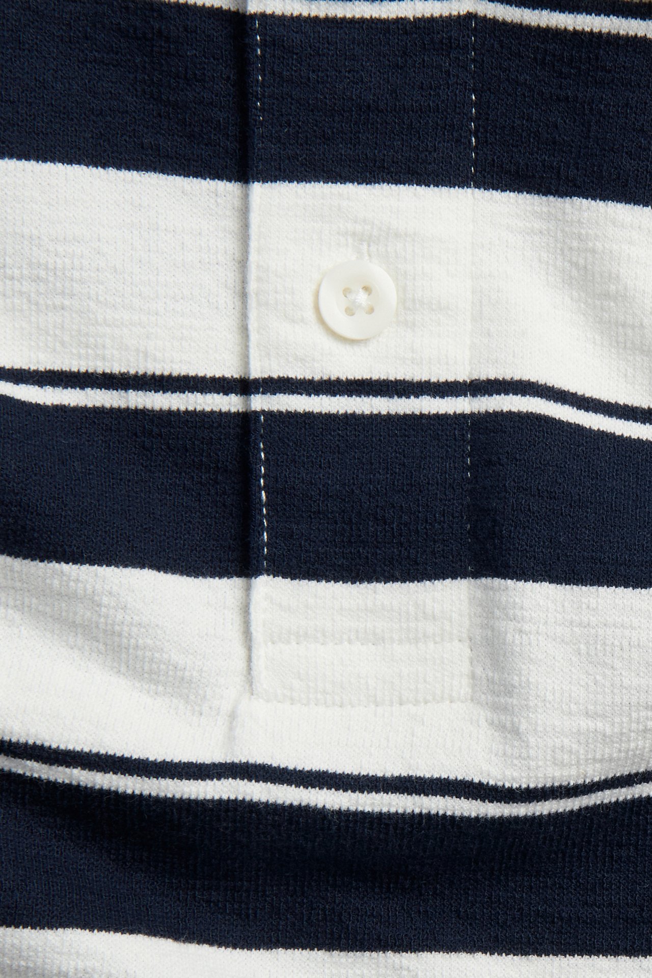 Lähikuva tummansinisestä ja valkoisesta raidallisesta puuvillapikeepaidasta valkoisella napilla.