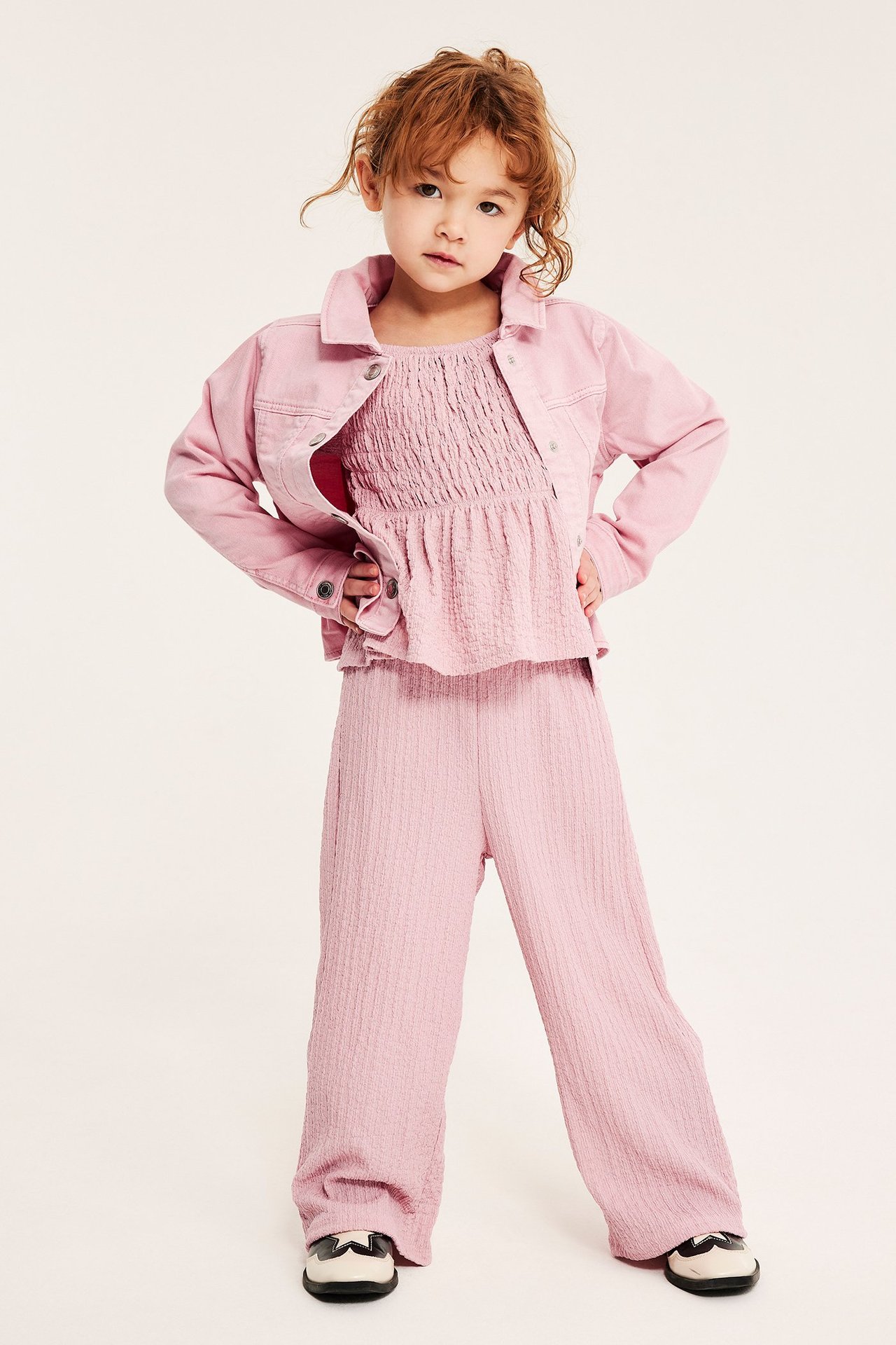 Från framsidan: ett barn i ljusrosa krinklade culottebyxor, en smocktopp och en öppen jeansjacka.