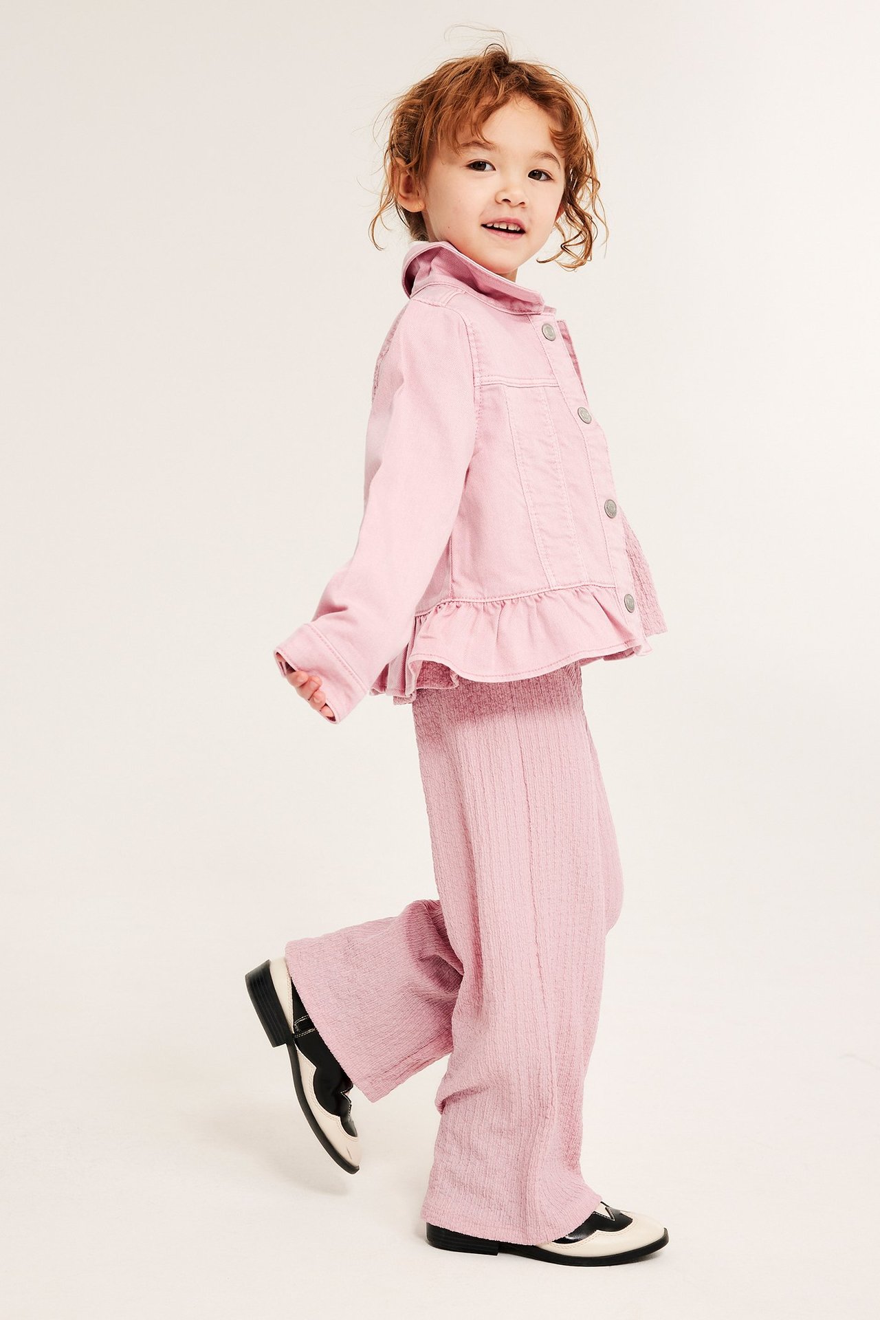 Widok z boku dziecka w jasnoróżowych, marszczonych kulotach, pasującej kurtce i dwukolorowych butach.