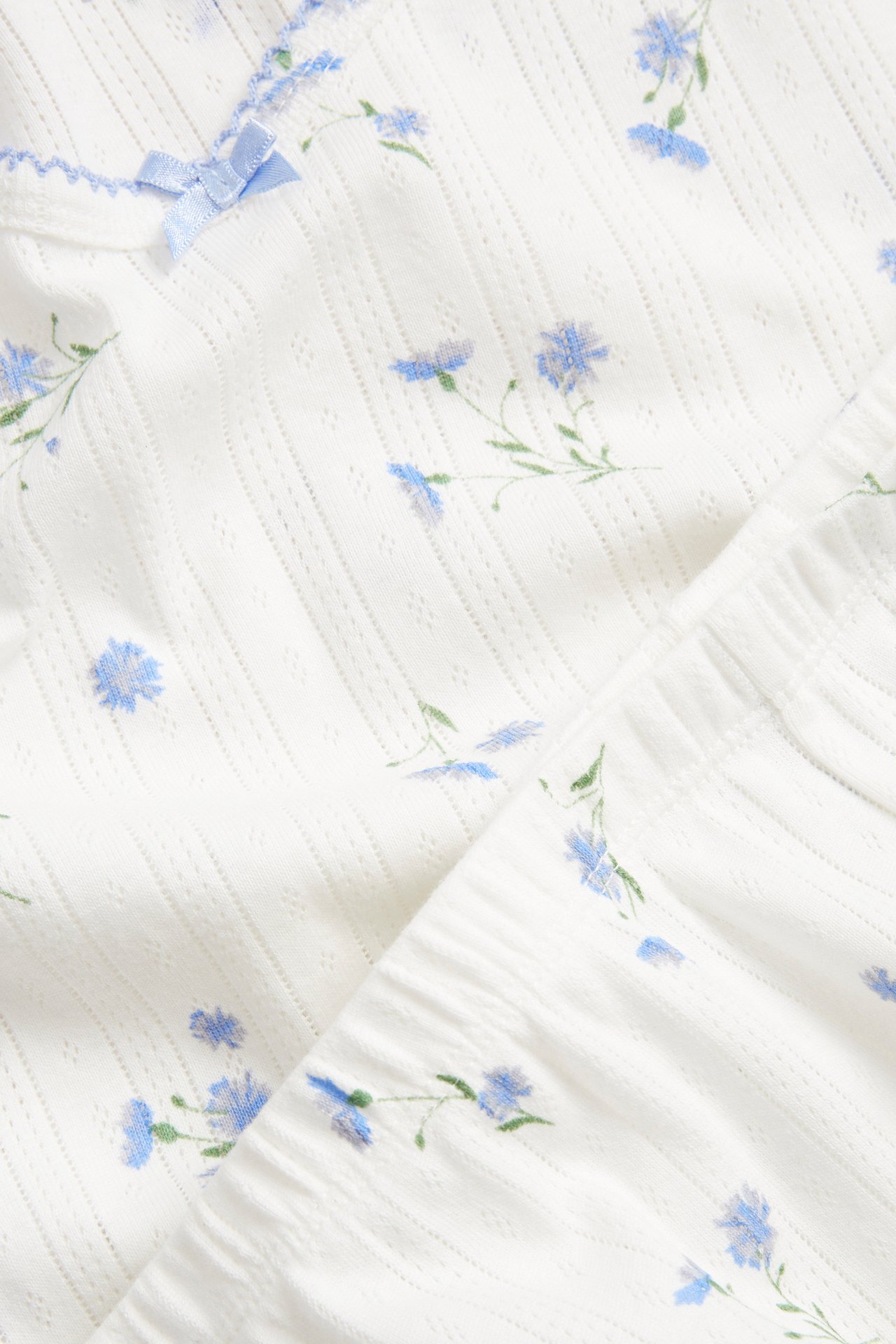 Zbliżenie na biały zestaw piżamowy z bawełny pointelle w jasnoniebieski kwiatowy wzór, z małą kokardą i koronką.