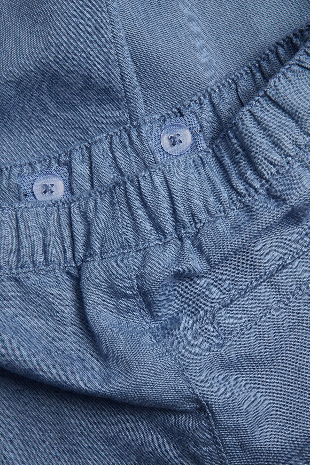 Lähikuva sinisistä pellavasekoitehousuista, joissa joustava vyötärö ja säädettävät nappisäätimet.