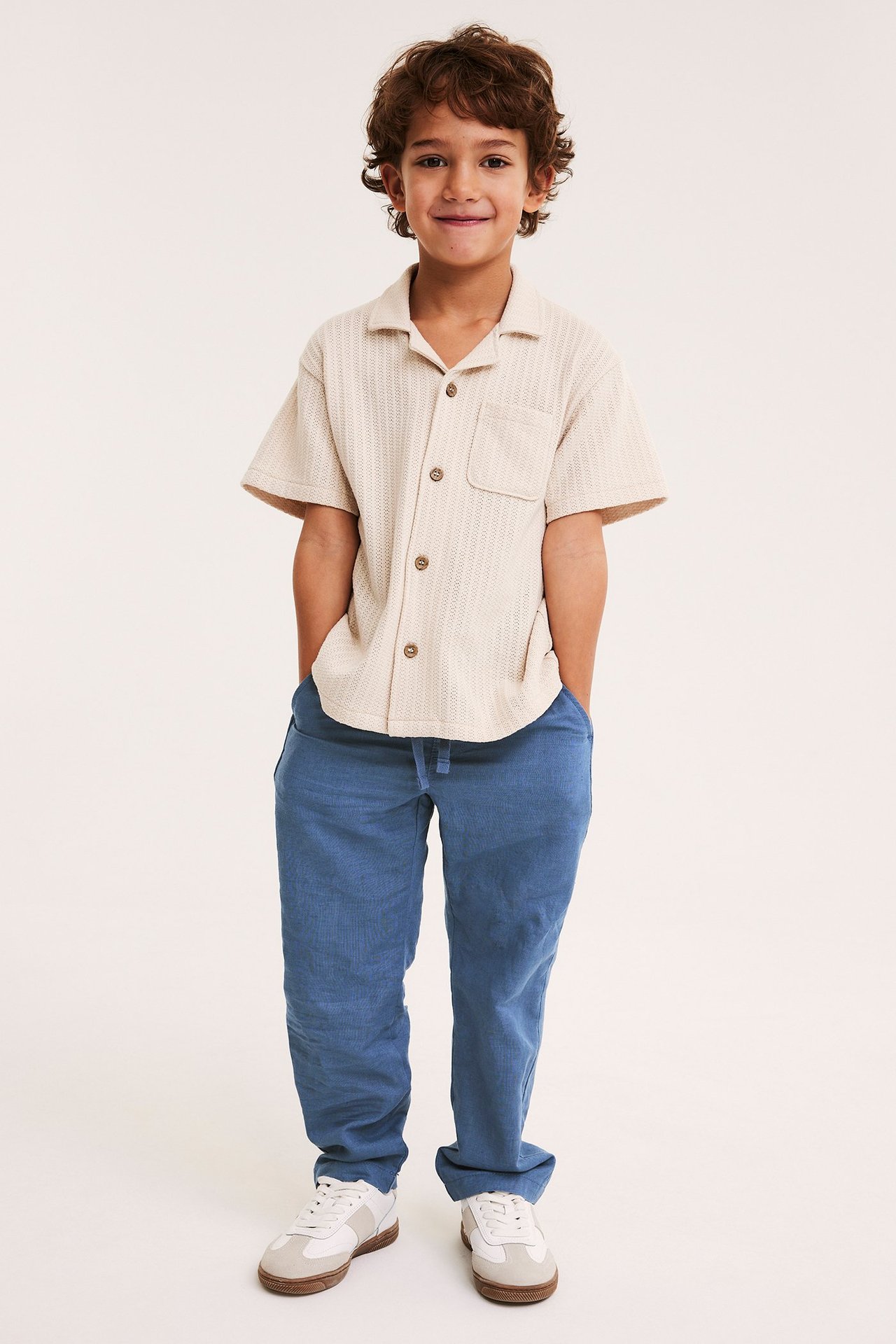 Edestä päin: lapsi yllään siniset pellavasekoitehousut, beige paita ja valkoiset tennarit.