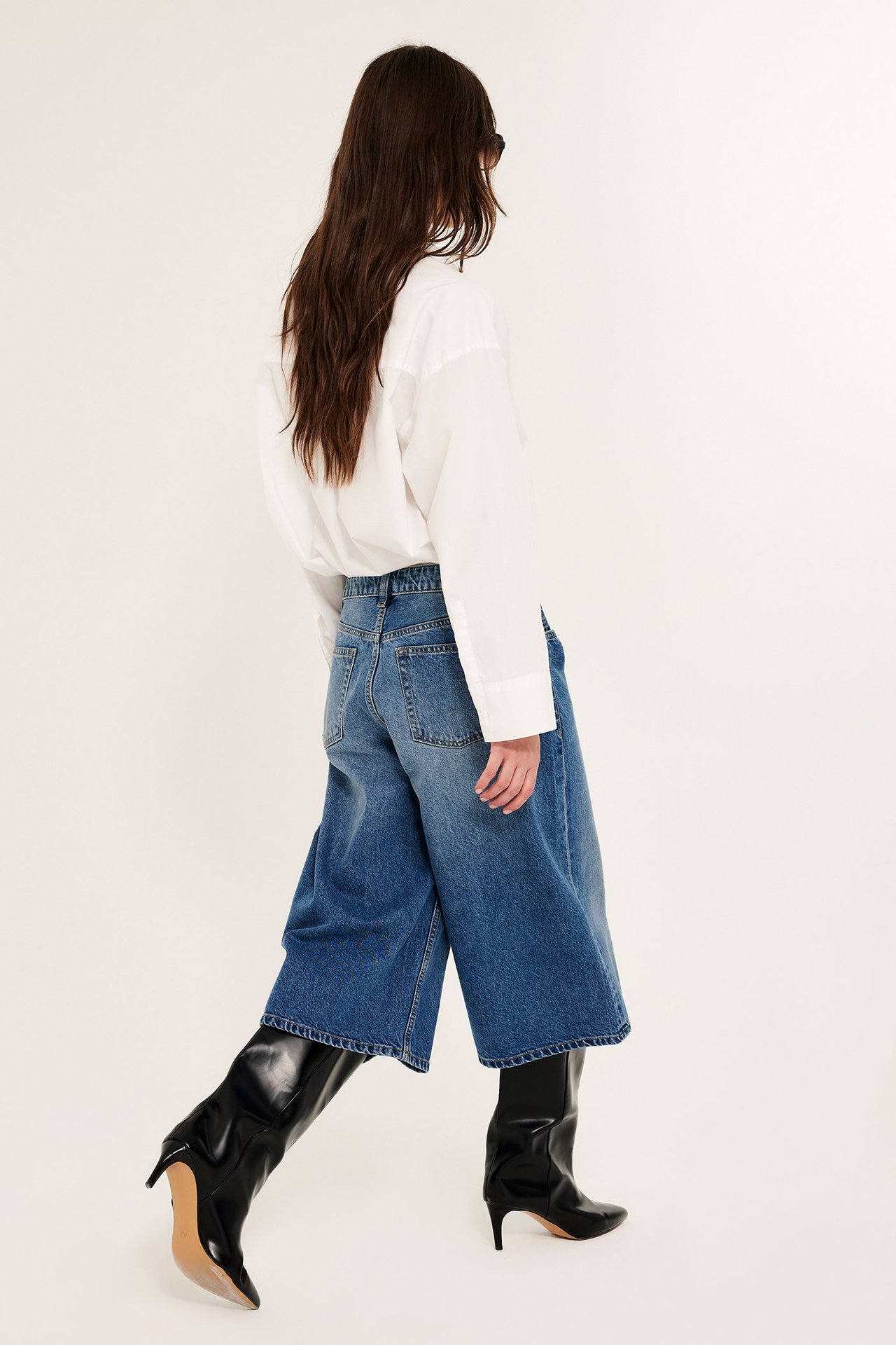 Takakuva naisesta väljissä sinisissä denim bermuda-shortseissa, valkoisessa paidassa ja mustissa saappaissa.