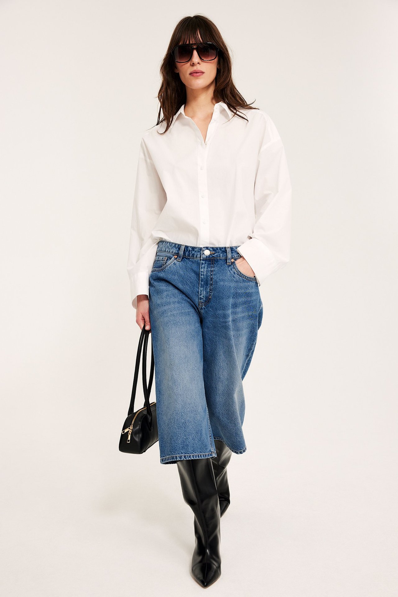 Nainen sinisissä denim-bermudashortseissa, valkoinen paita, mustat saappaat, aurinkolasit, musta laukku. Edestä.