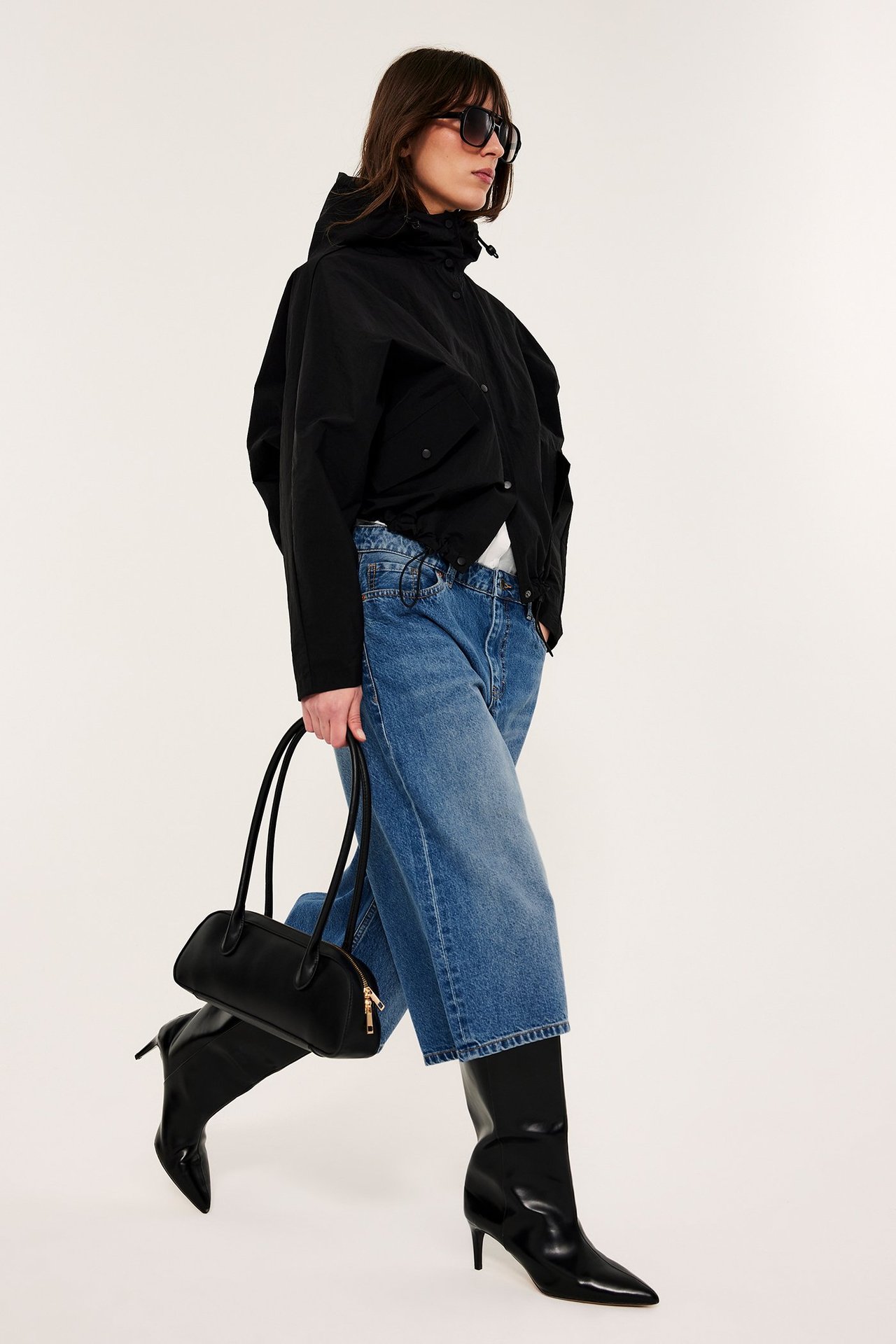 Naisen sivukuva: musta hupullinen takki, siniset leveälahkeiset farkut, mustat saappaat, musta käsilaukku.