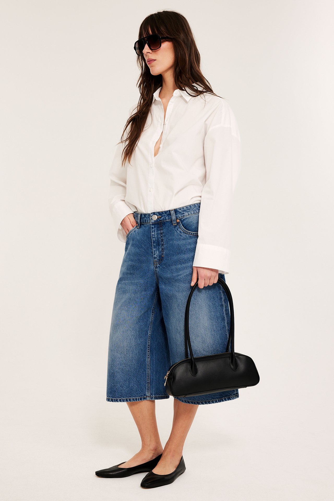 Sivukuva: nainen pukeutuu leveisiin sinisiin denim-sortseihin, valkoiseen paitaan, mustiin kenkiin, aurinkolaseihin, pitelee mustaa käsilaukkua.
