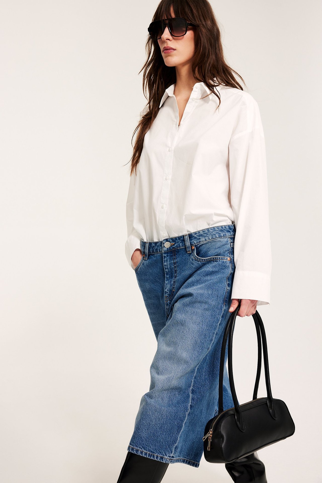 Edestä: Nainen rentoissa, leveissä sinisissä denimsortseissa, valkoisessa paidassa, mustissa aurinkolaseissa, pitäen mustaa käsilaukkua.