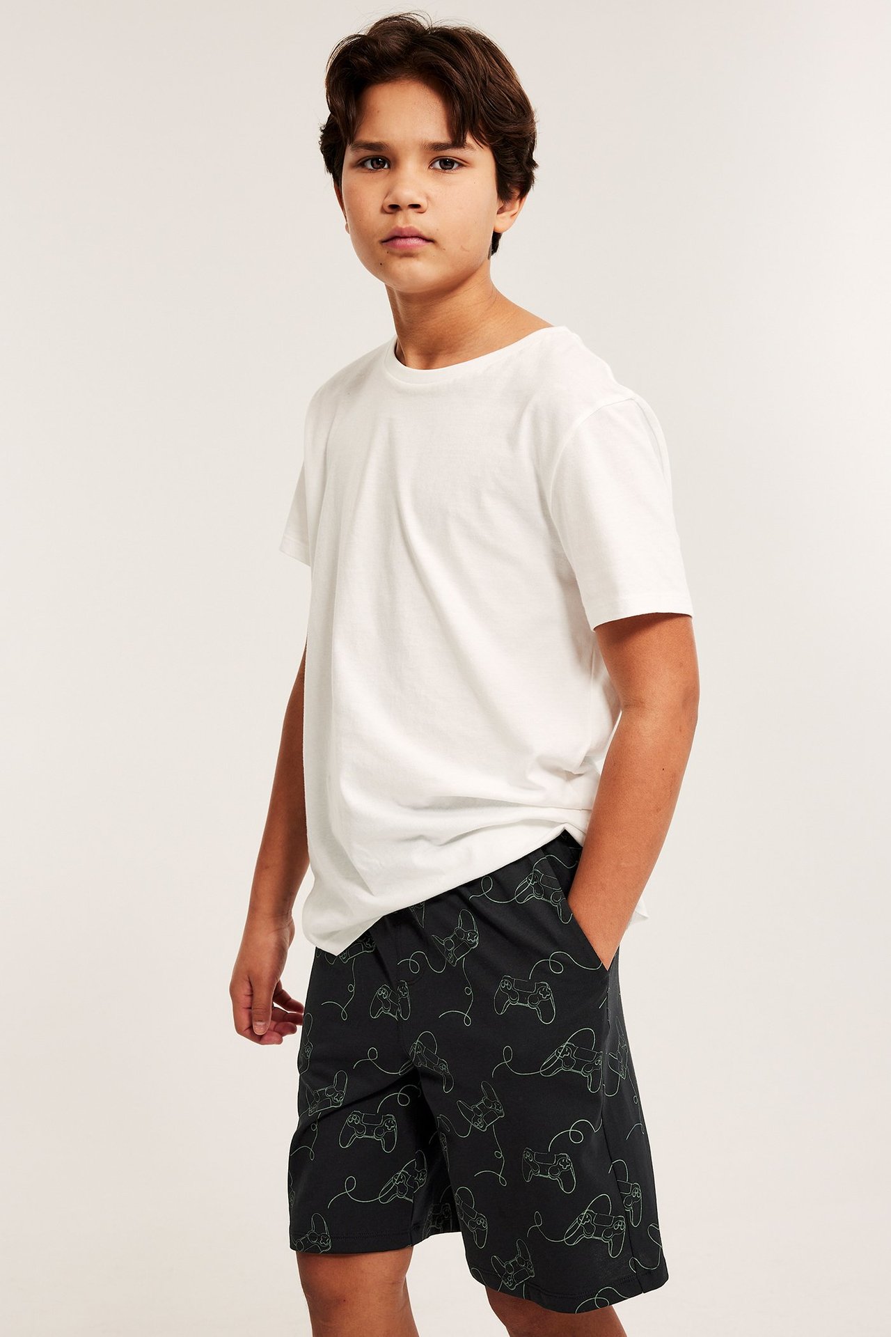 Edestäpäin: Lapsi mustissa pyjamashortseissa, joissa vihreä peliohjainkuvio, ja valkoisessa puuvilla-T-paidassa.