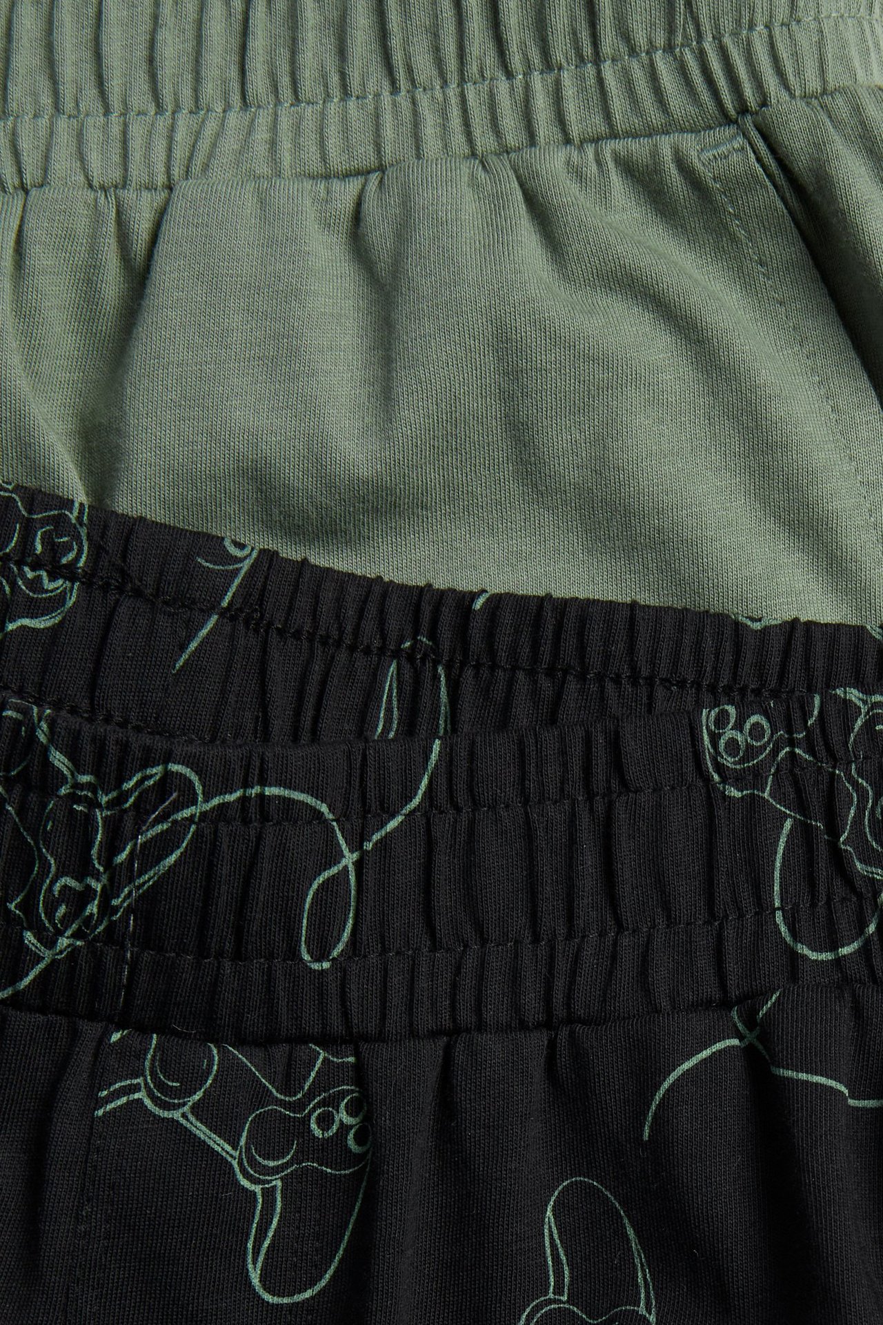Närbild av bomulls-pyjamasshorts 2-pack: ett svart med ljusgrönt mönster, ett enfärgat olivgrönt.