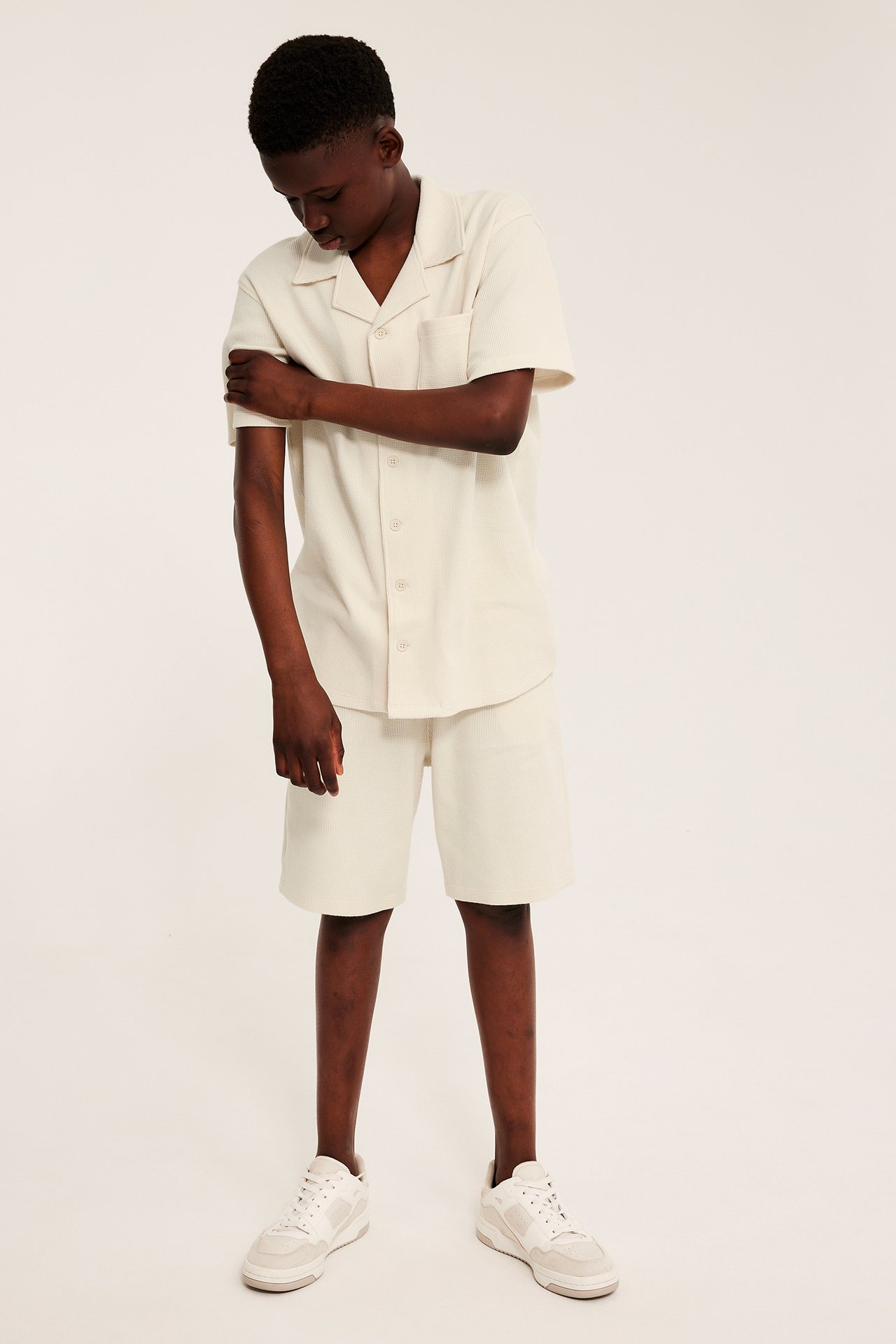 Lapsi edestäpäin: vaaleanbeige vohvelipuuvillainen lyhythihainen paita, shortsit ja valkoiset tennarit.