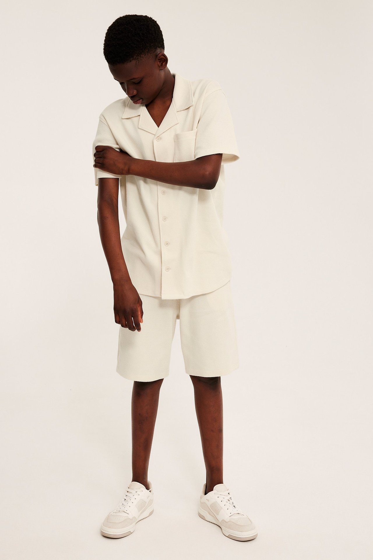 Ett barn i ljusbeige våfflad kortärmad skjorta, matchande shorts och vita sneakers, framifrån.