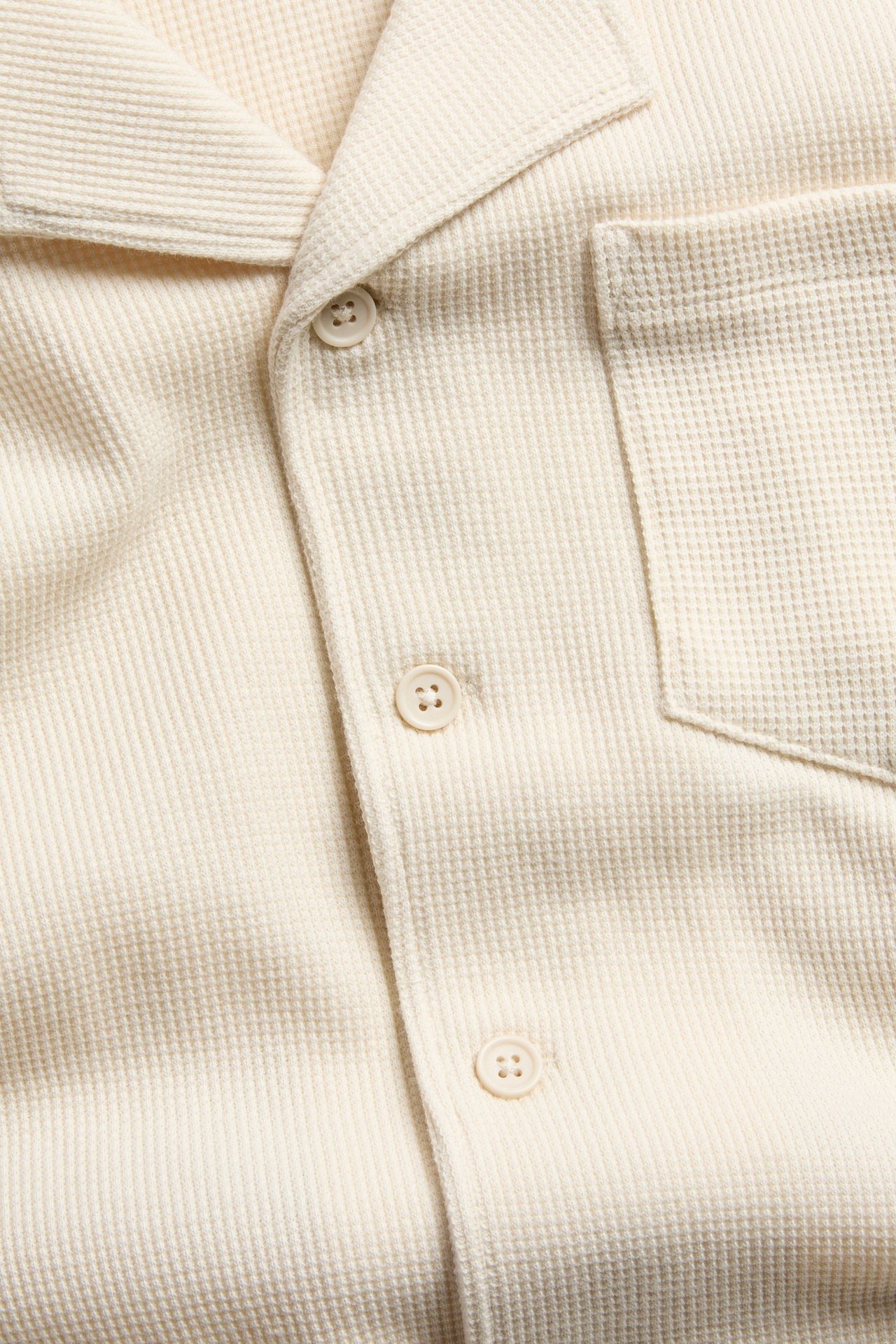 Zbliżenie na jasnobeżową koszulę bawełnianą w wafel, z kołnierzem resortowym, guzikami i kieszenią.