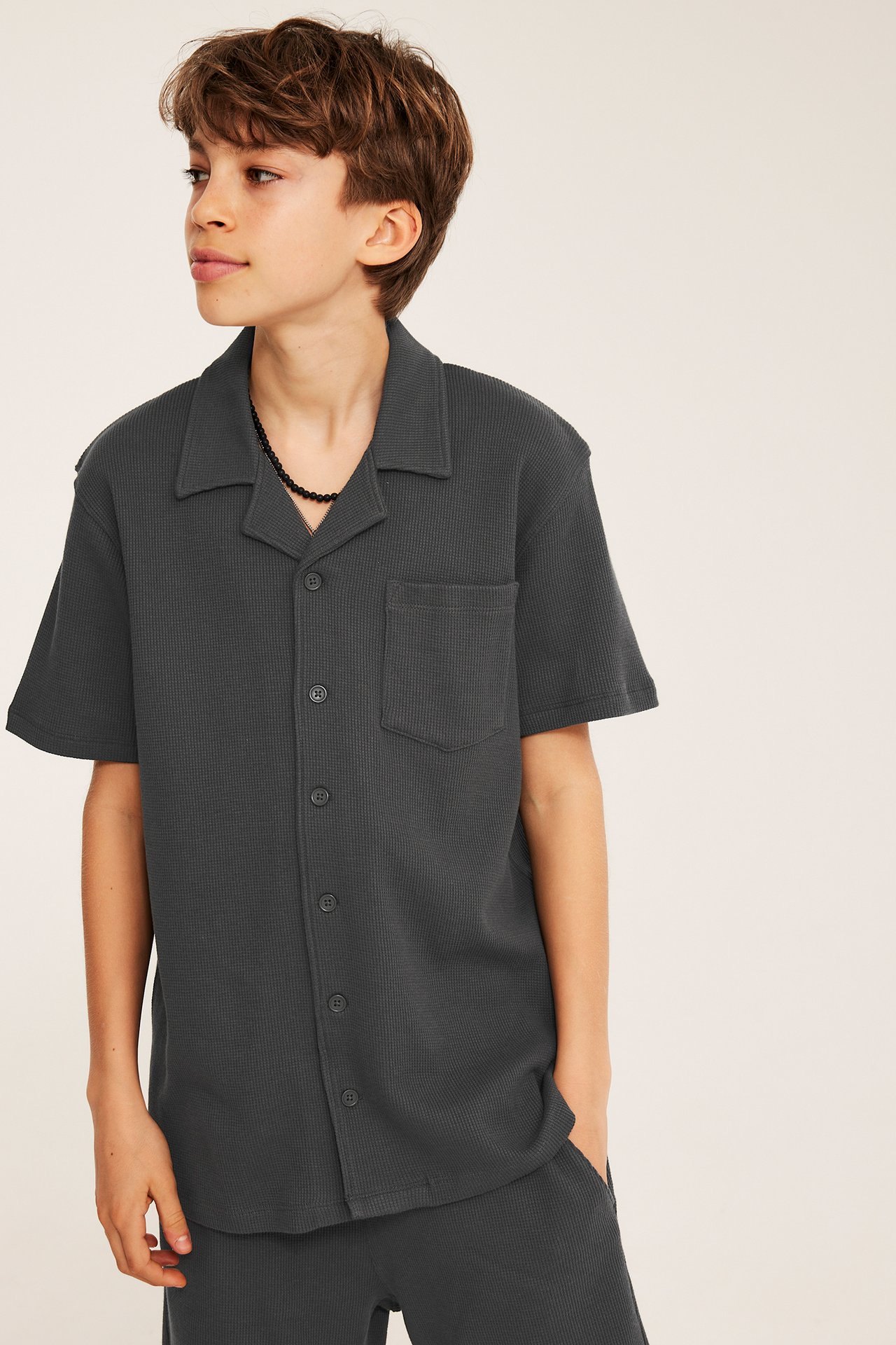 Edestä: Lapsi tummanharmaassa lyhythihaisessa vohvelikankaisessa puuvillapaidassa, yhteensopivat shortsit, musta kaulakoru.