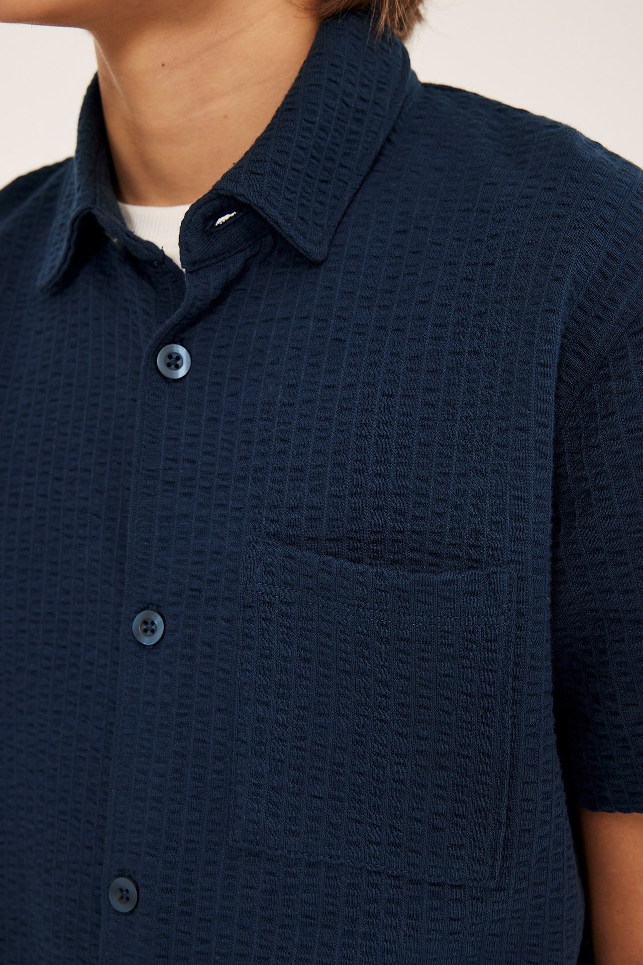 Zbliżenie przód: Granatowa koszula dziecięca z krótkim rękawem seersucker, guziki, kieszeń, na białym T-shircie.