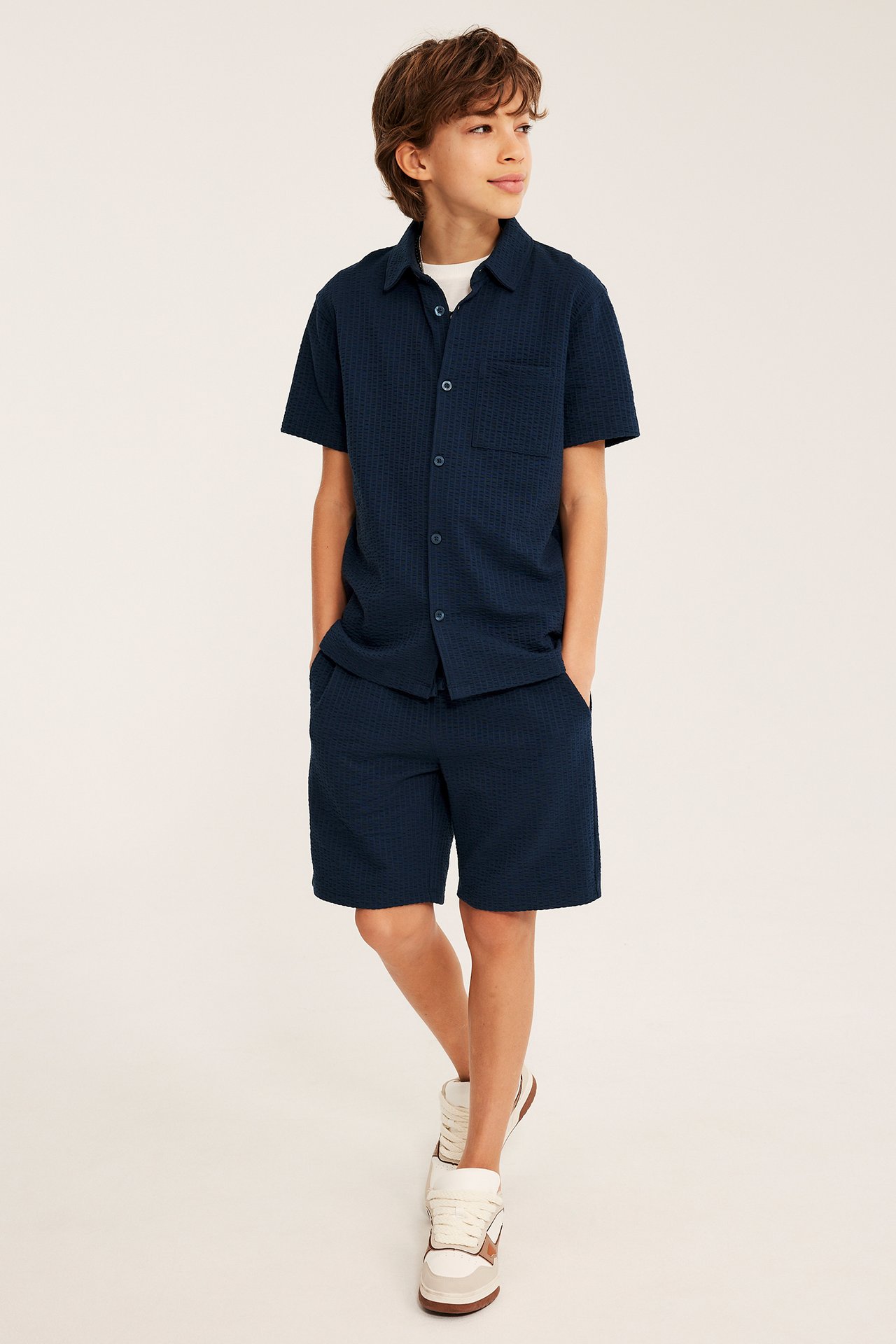 Edestä: Poika tummansinisessä lyhythihaisessa seersucker-paidassa valkoisen t-paidan päällä ja shortseissa.