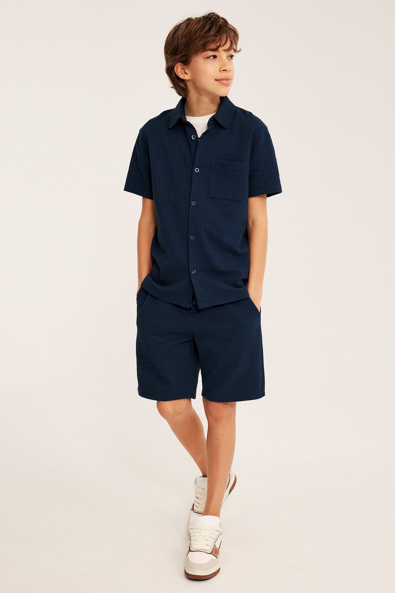 Widok z przodu: Chłopiec w granatowej koszuli z krótkim rękawem z seersuckeru, białym t-shircie i pasujących szortach.