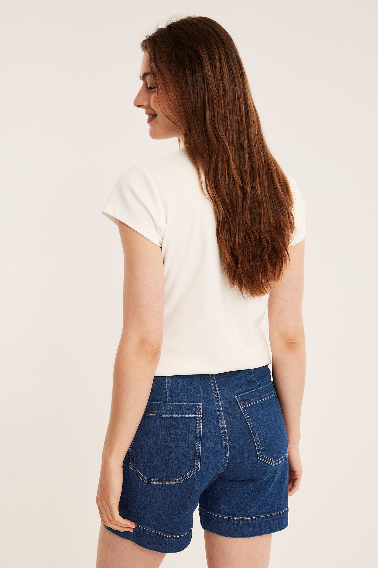 Takasivukuva naisesta tummansinisissä denimsortseissa, joissa paikkataskut, ja vaaleassa lyhythihaisessa paidassa.