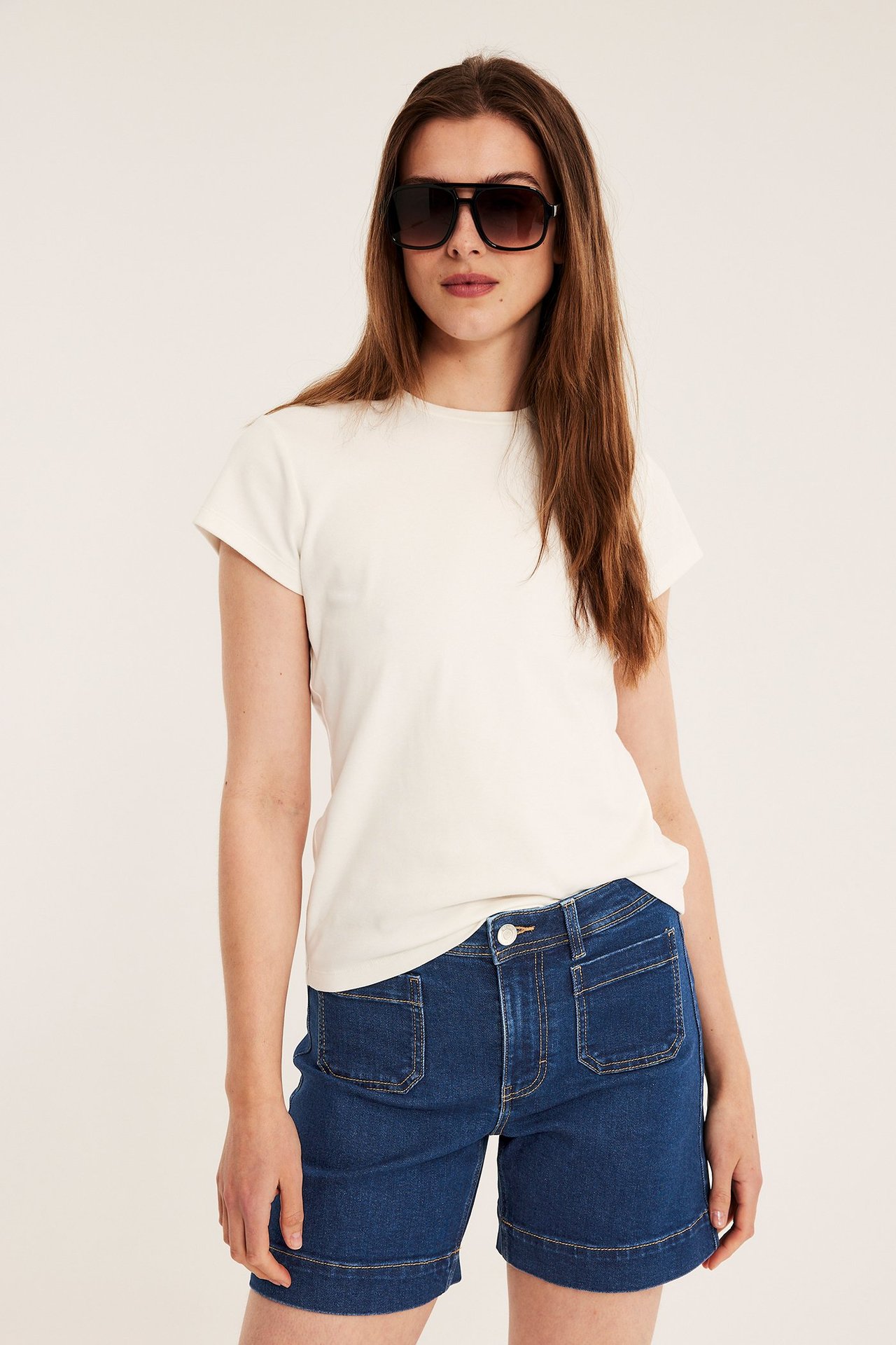 Edestä: Nainen sinisissä farkkushortseissa paikkutaskuilla, valkoisessa t-paidassa ja mustissa aurinkolaseissa.