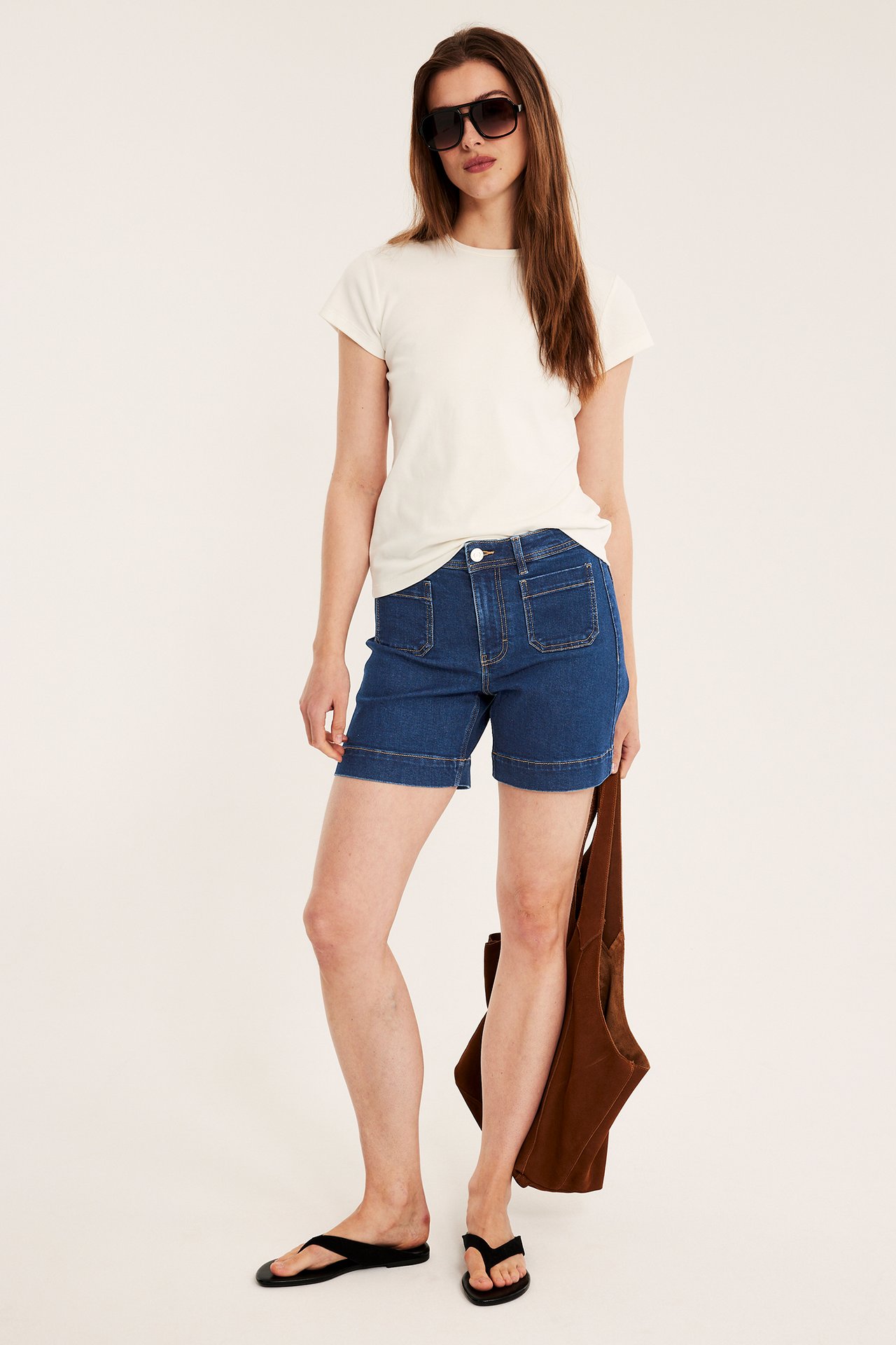 Nainen sinisissä paikkataskullisissa farkkushortseissa, luonnonvalkoisessa t-paidassa, aurinkolaseissa, varvassandaaleissa, ruskea laukku kädessä.