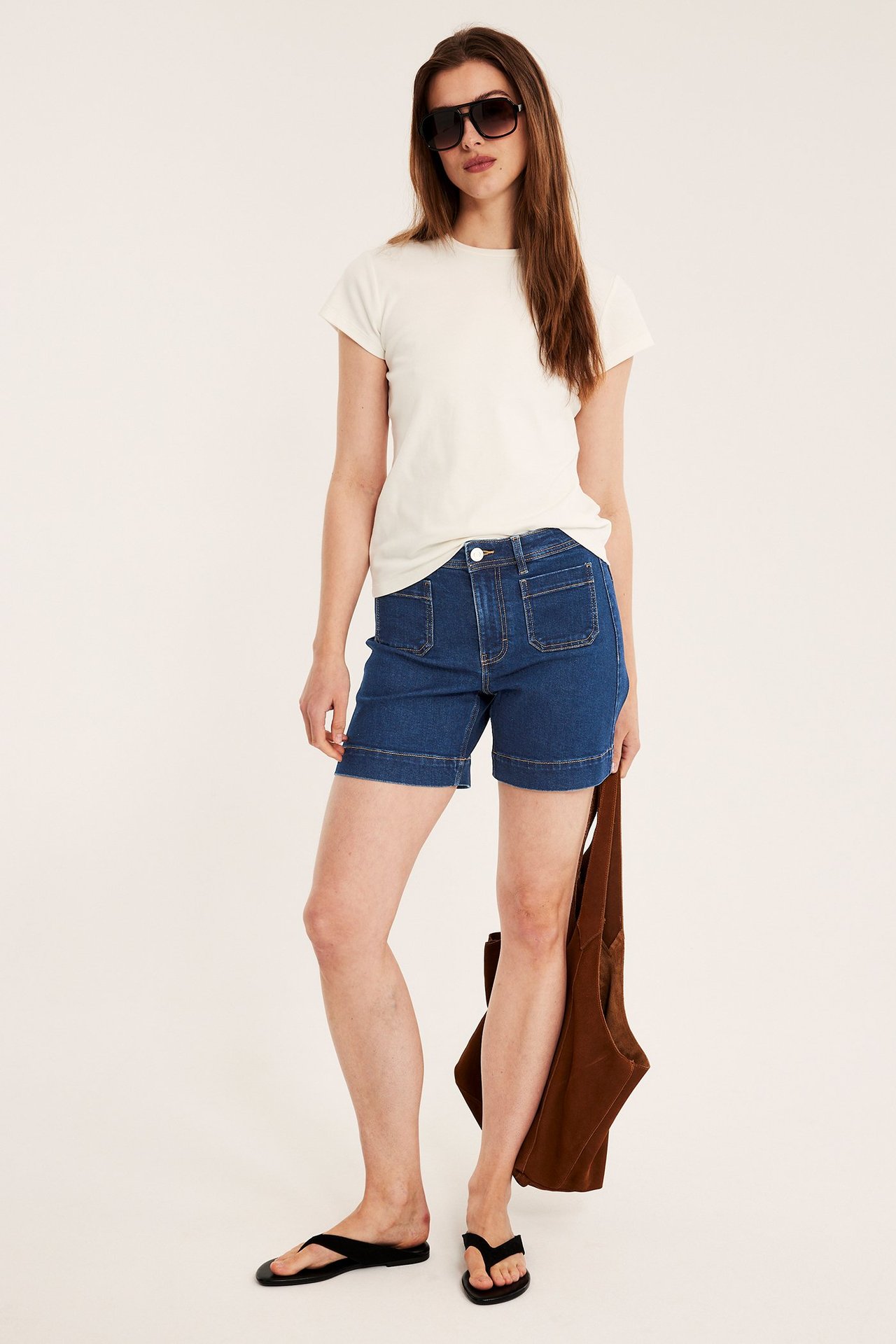 Nainen sinisissä paikkataskullisissa farkkushortseissa, luonnonvalkoisessa t-paidassa, aurinkolaseissa, varvassandaaleissa, ruskea laukku kädessä.