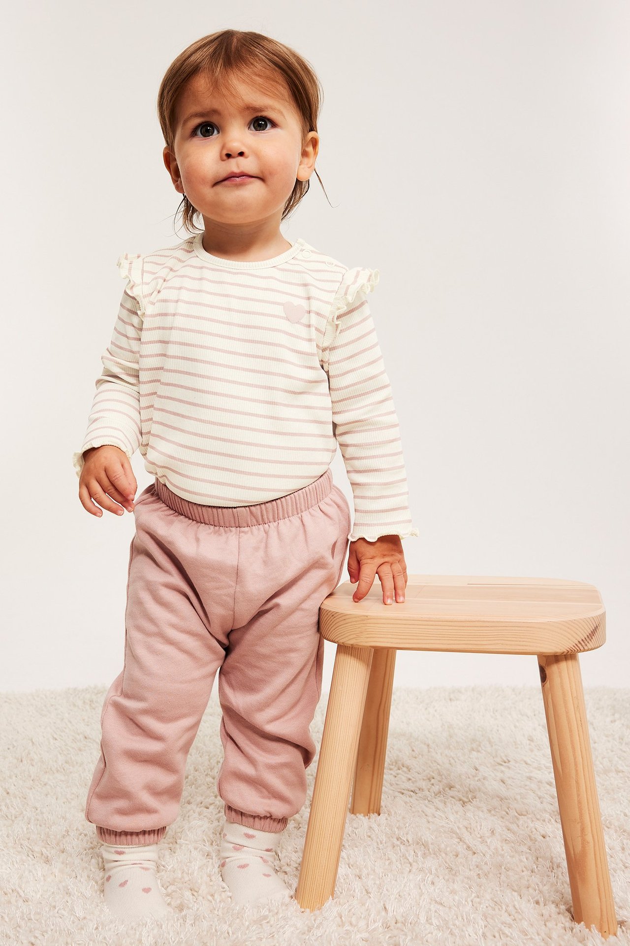 Widok z przodu: Niemowlę w prążkowanym topie w paski z falbanami, różowym sercem, różowymi spodniami i białymi skarpetkami w serca.