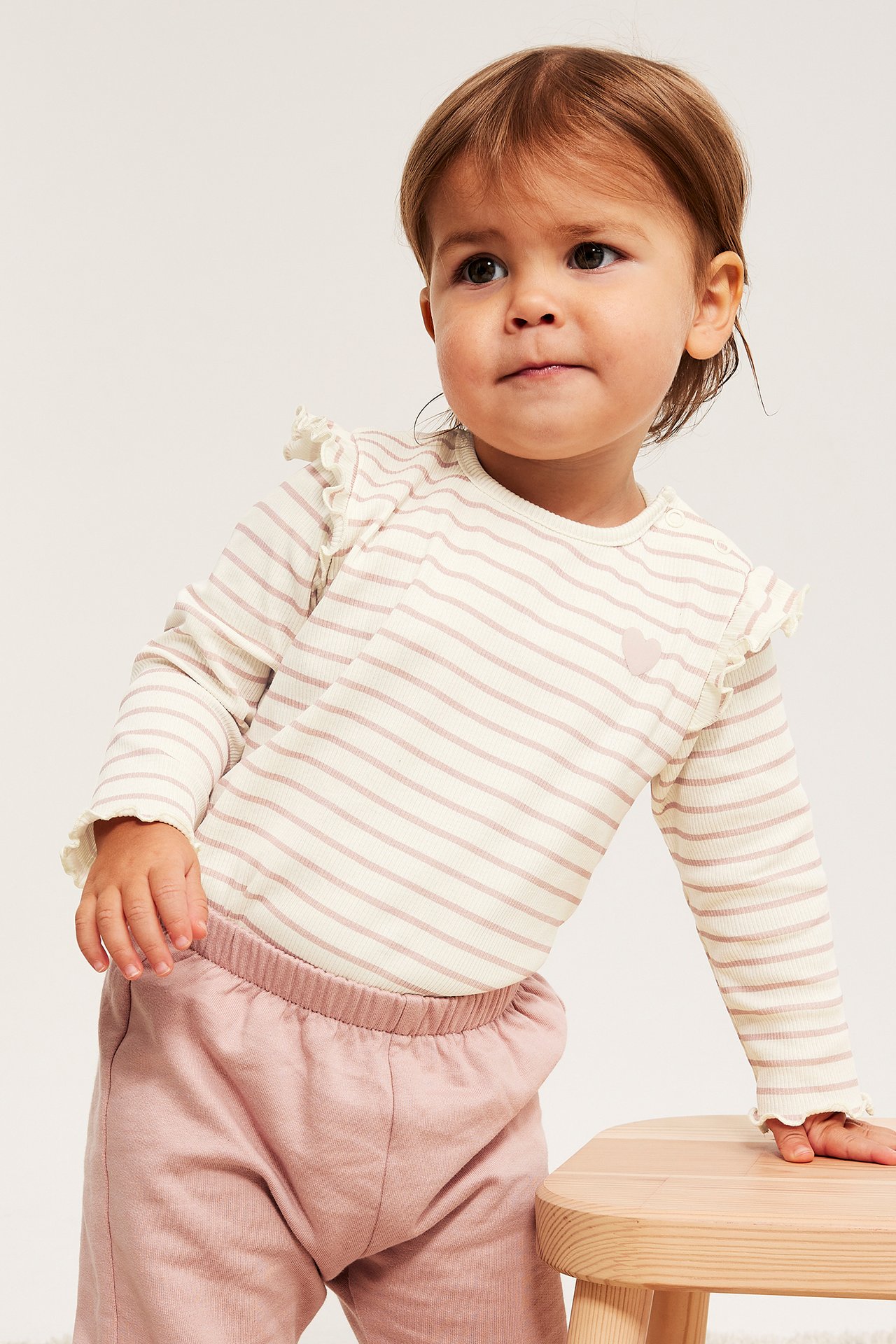 Niemowlę, widok z przodu, w kremowym prążkowanym topie z długim rękawem, różowymi paskami, falbankami i różowych dresach.