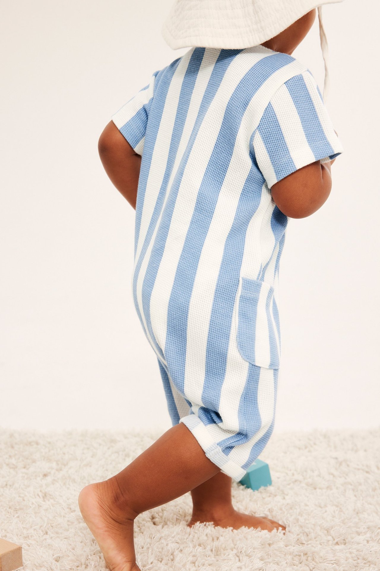 Sidovy av baby i ljusblå och vitrandig våffelromper med ficka och vit solhatt.