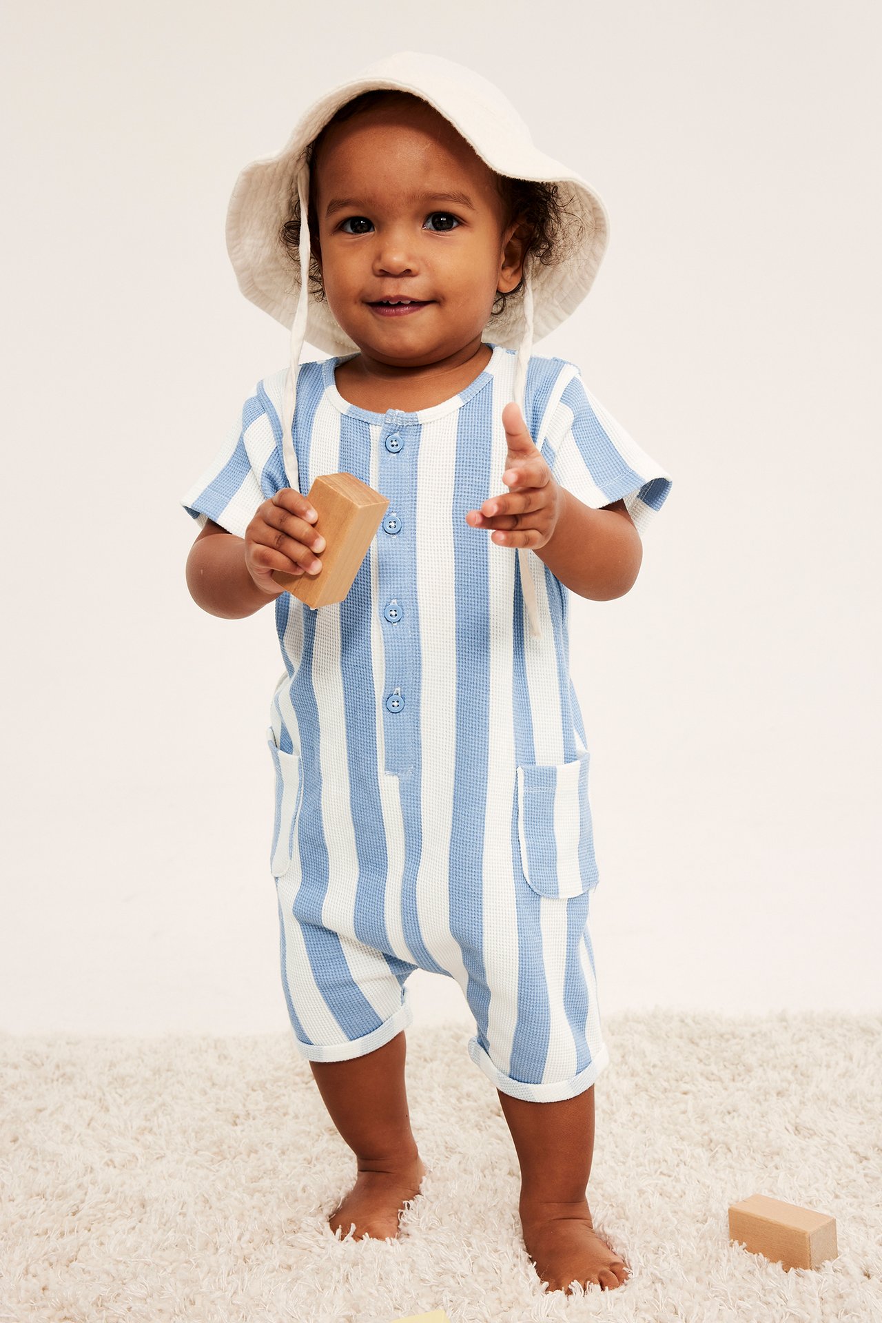 Framsida: Baby i ljusblå/vit randig våfflad romper med knappar, fickor, vit solhatt, håller i en kloss.