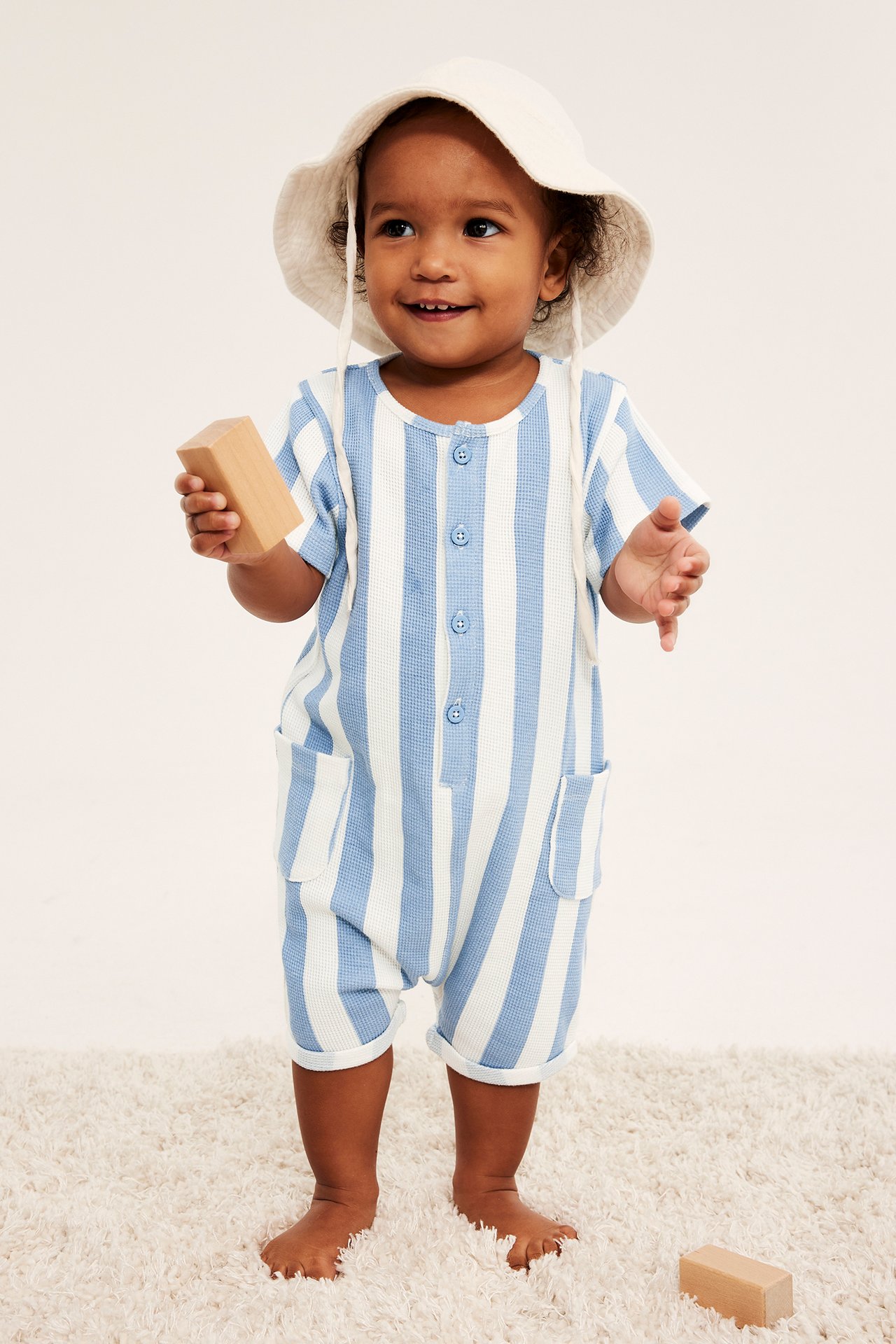 Baby bär en randig ljusblå och vit våffelromper med knappar, fickor och en beige hatt.