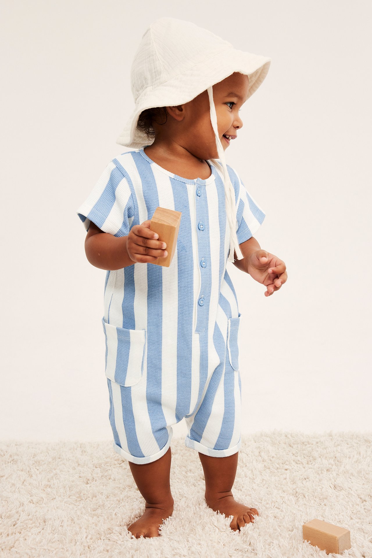 Baby i ljusblå, vitrandig våfflad romper med knappar, fickor och krämvit solhatt, håller i kloss.
