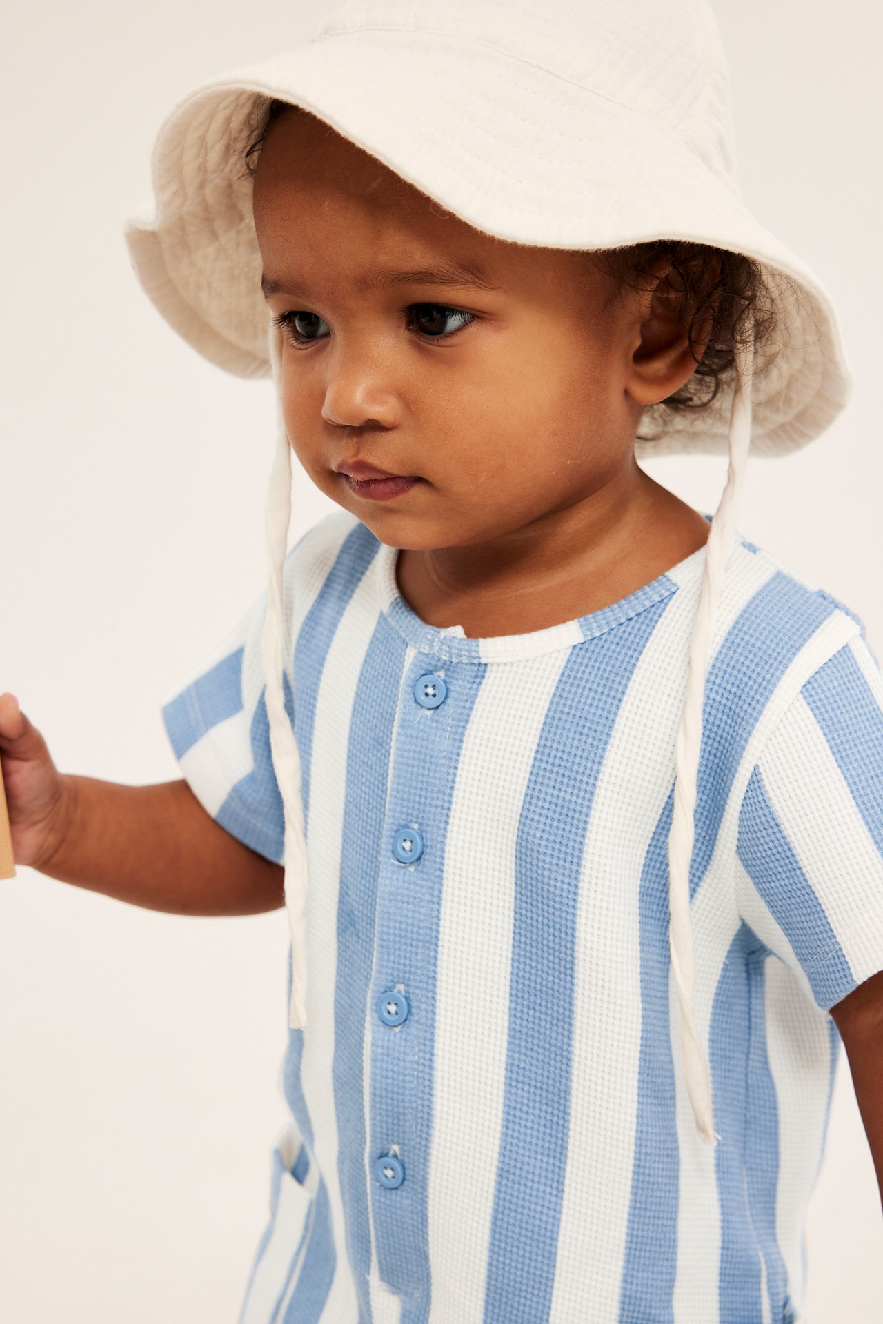 Närbild på baby i ljusblå/vit randig våfflad romper med knappar och en cremefärgad hatt.