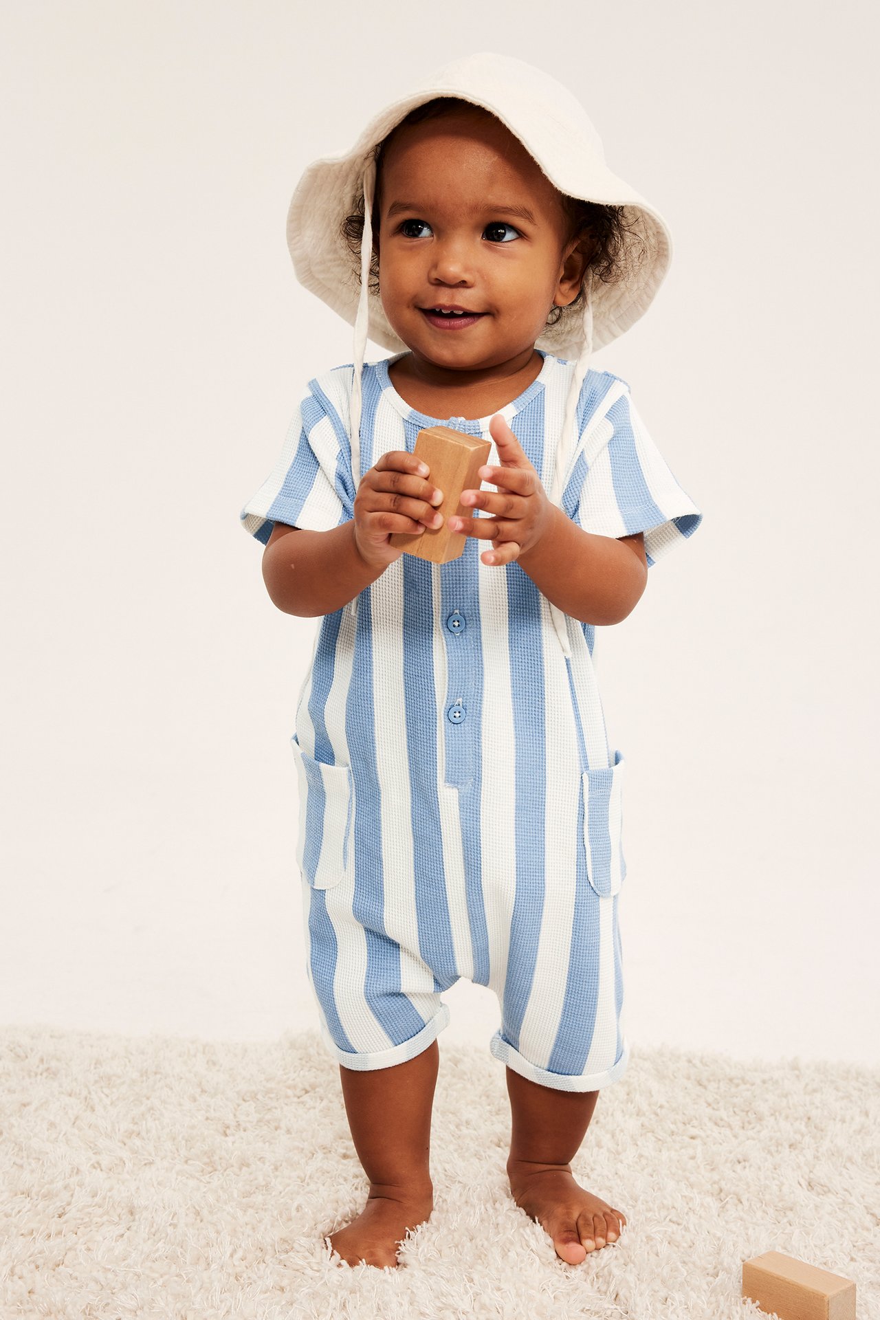 Baby i ljusblå och vitrandig våfflad romper med knappar, fickor och en beige solhatt.