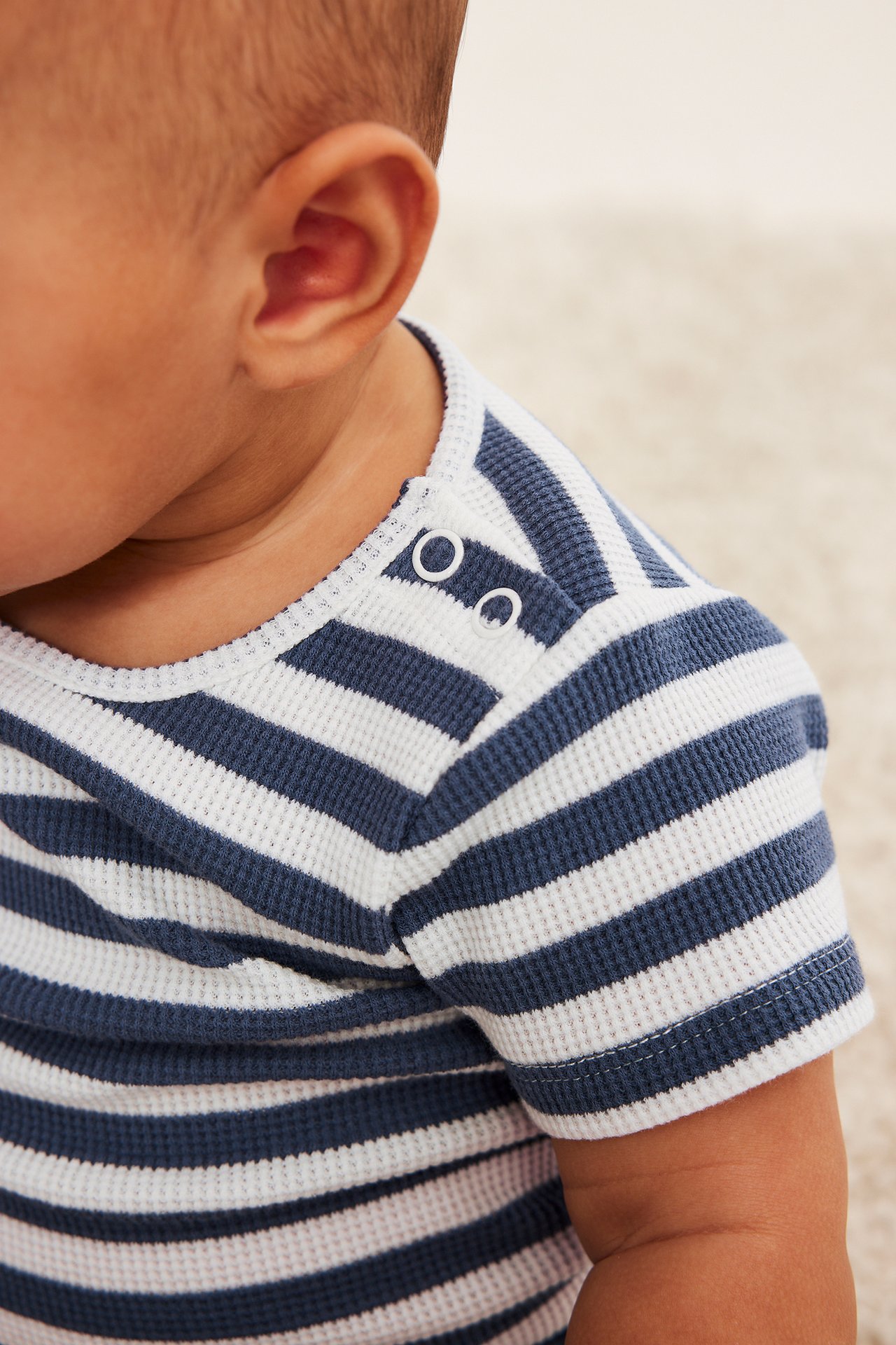 Närbild på baby som bär en blå- och vitrandig kortärmad våffelbody med vita tryckknappar på axeln.