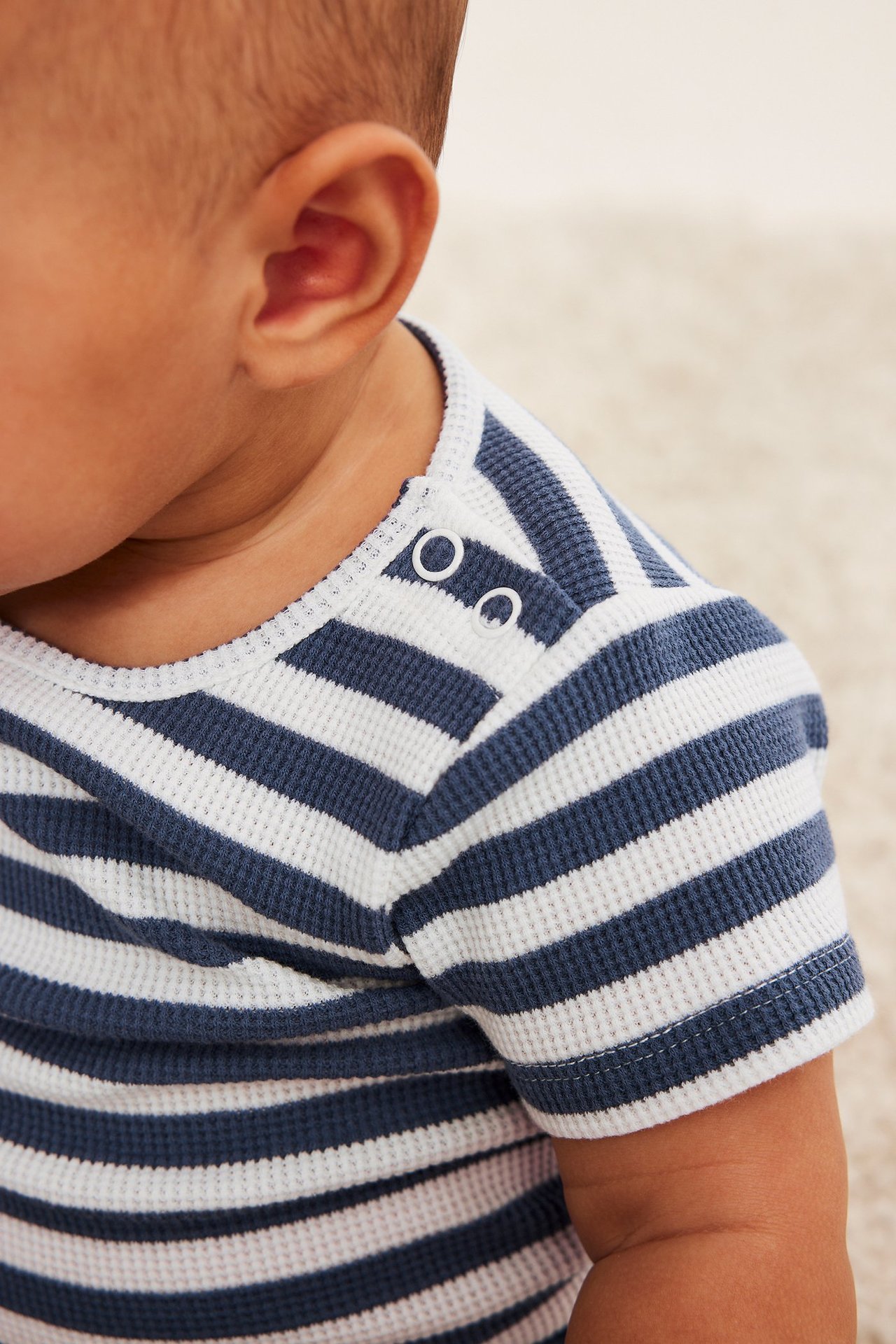 Lähikuva vauvasta, jolla on sinivalkoraidallinen lyhythihainen vohvelibody valkoisilla olkapainonapeilla.