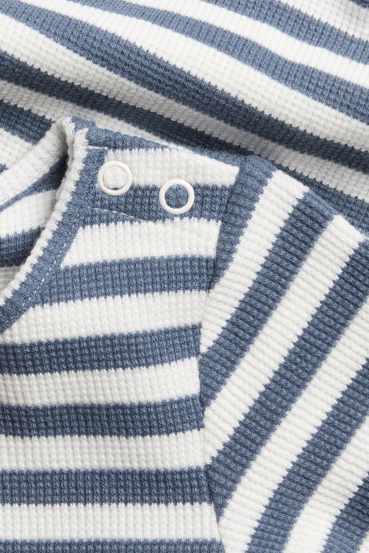 Nærbilde av en babybody i blå- og hvitstripete vaffelstrikk med hvite trykknapper på skulderen.