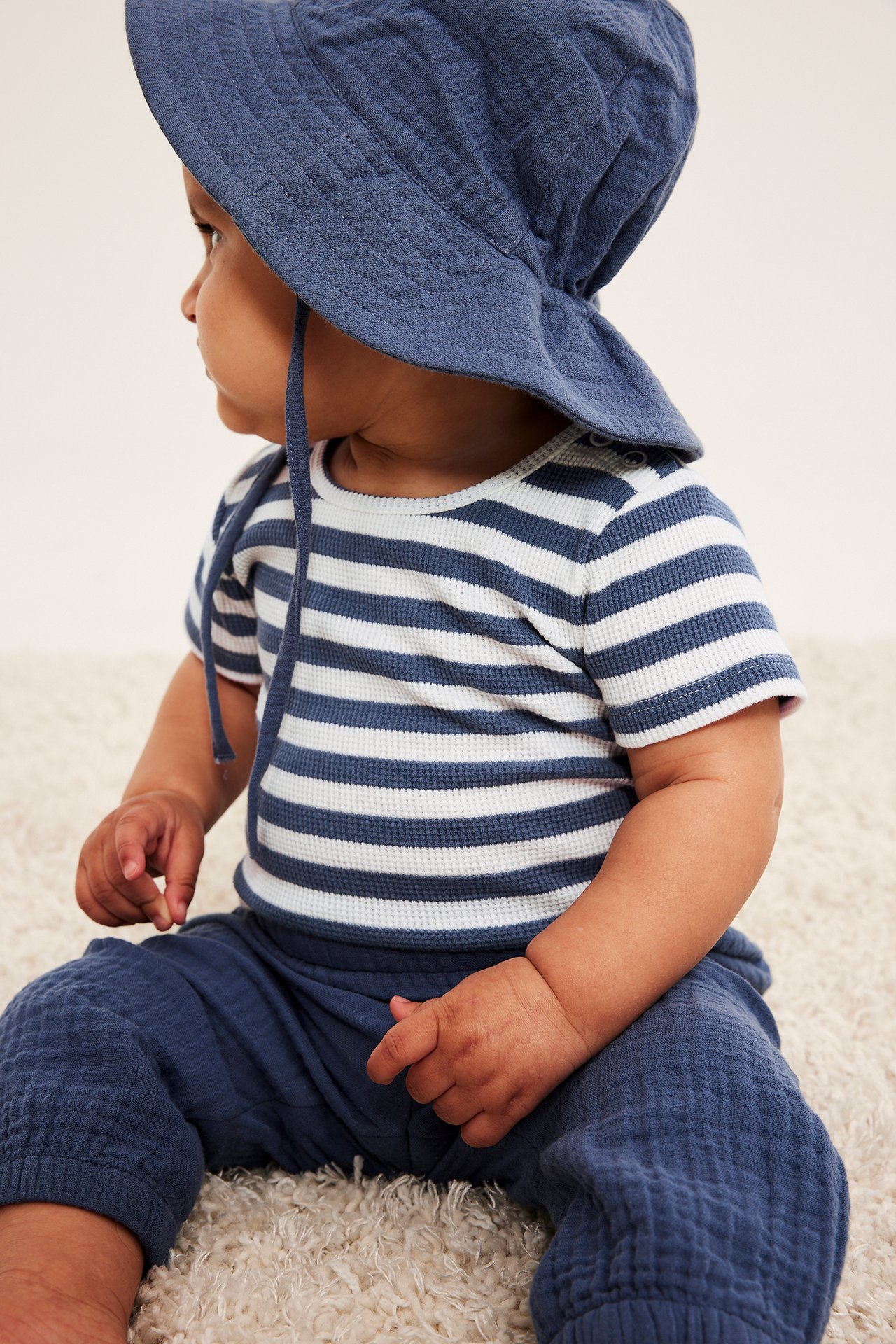 Baby, sidovy: en blåvitrandig våffelbody, blå våffelbyxor och en blå solhatt.