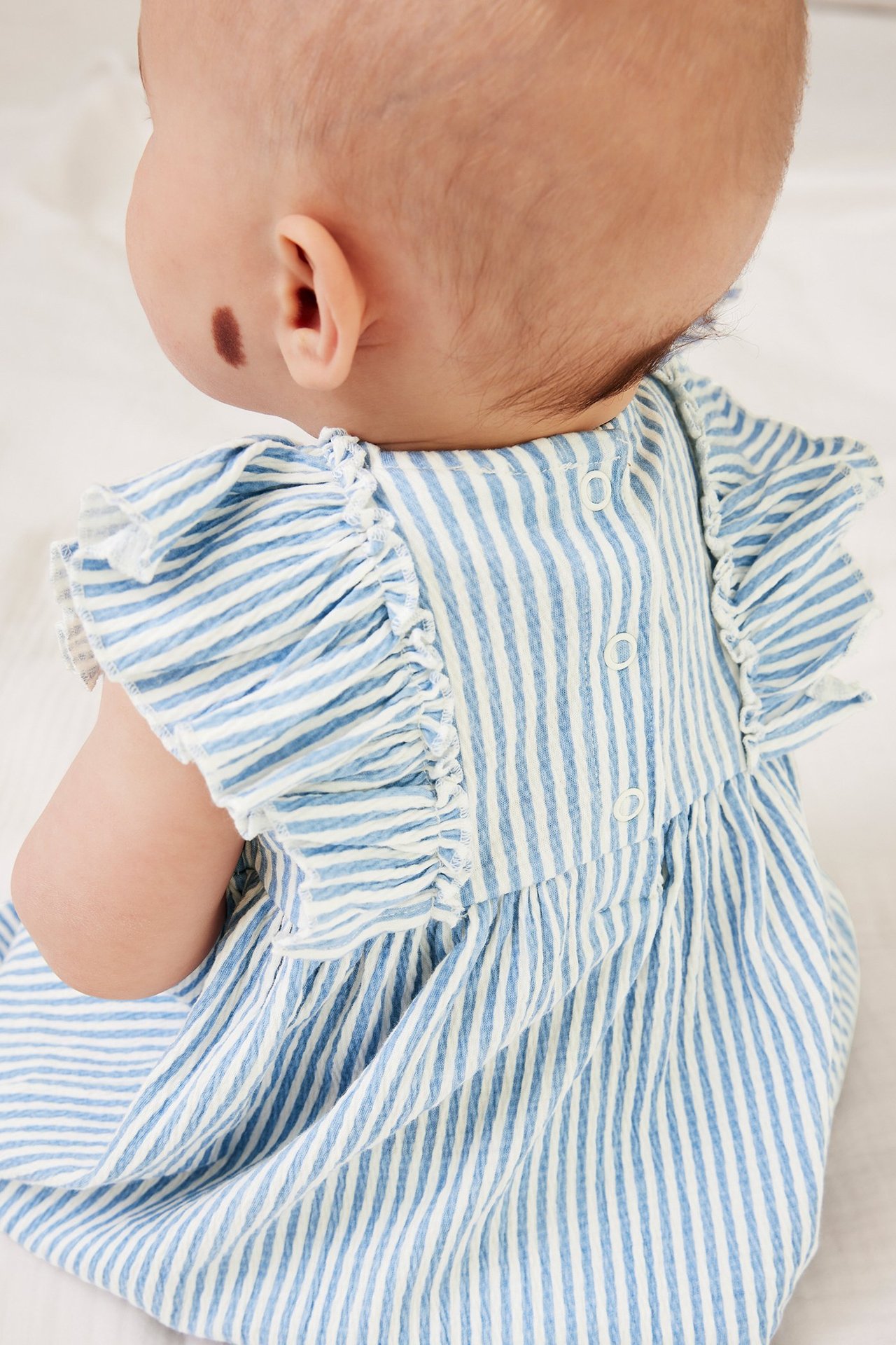 Takaapäin näkymä vauvasta vaaleansinivalkoraidallisessa jerseymekossa, jossa on olkaröyhelöt ja nepparit.