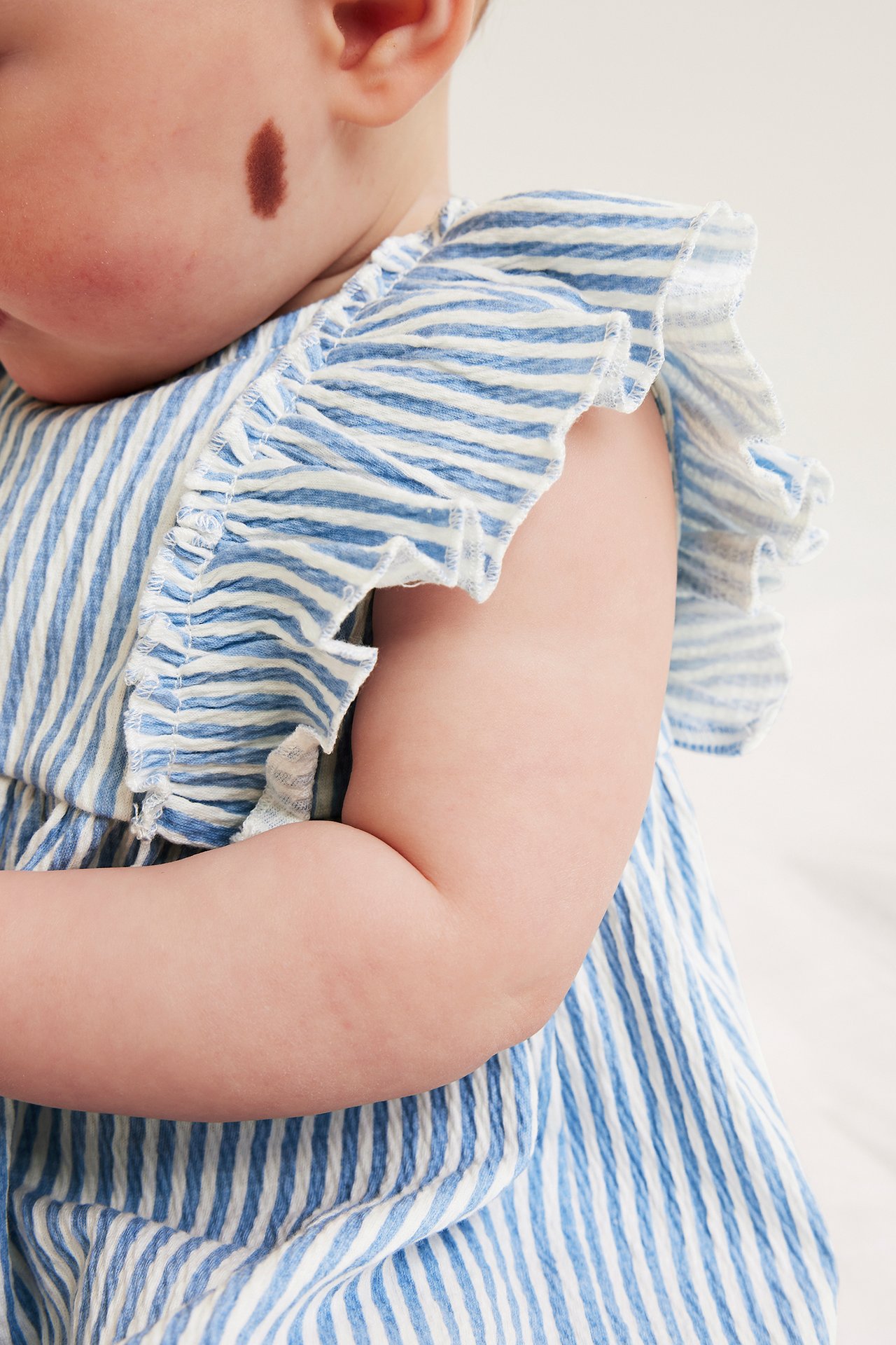 Lähikuva vauvasta, jolla on vaaleansinivalkoinen raidallinen kreppimekko röyhelöolkapäillä.