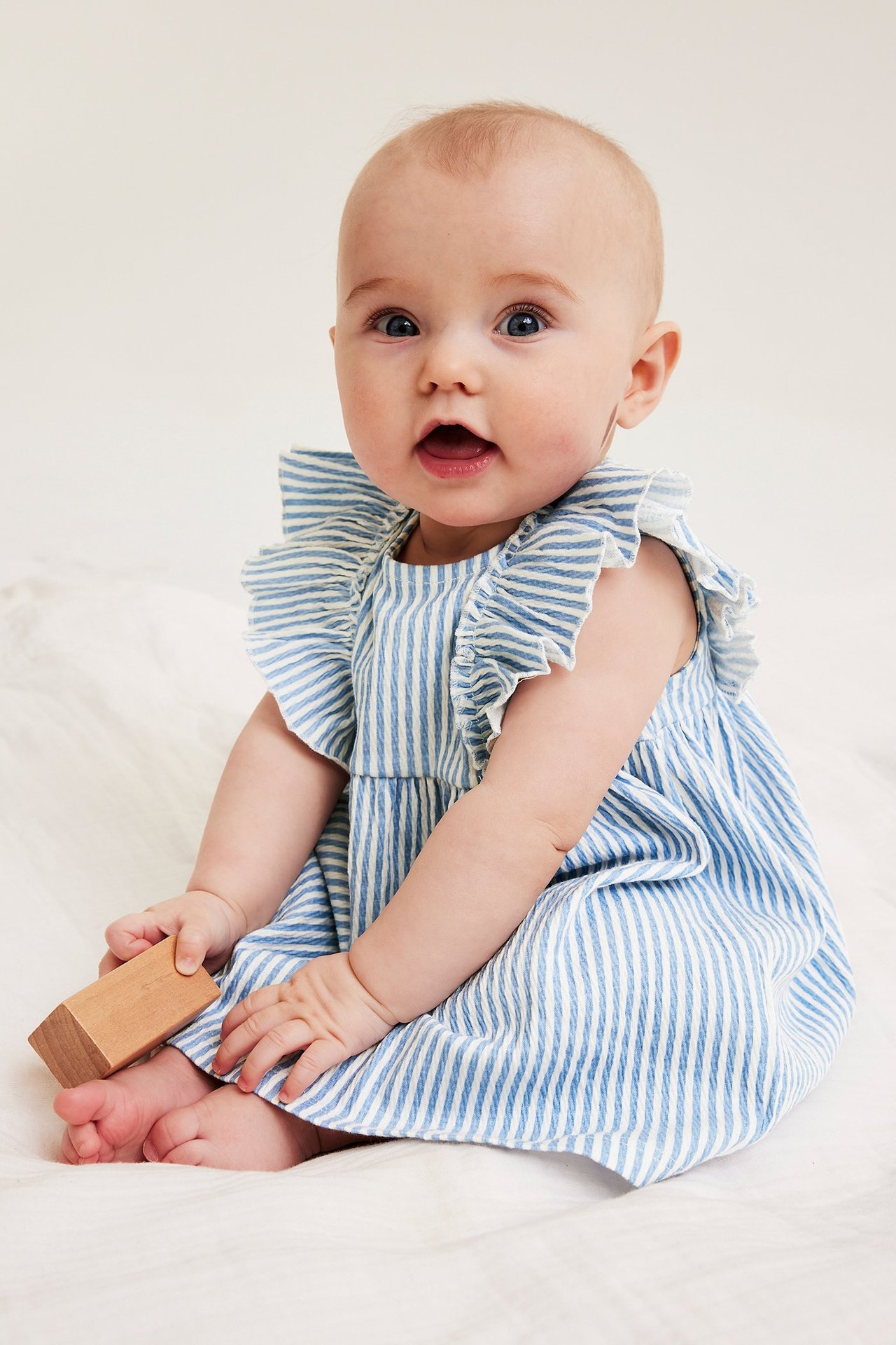 Widok z przodu dziecka w jasnoniebiesko-białej sukience dżersejowej w paski z falbanami, trzymającego drewniany klocek.