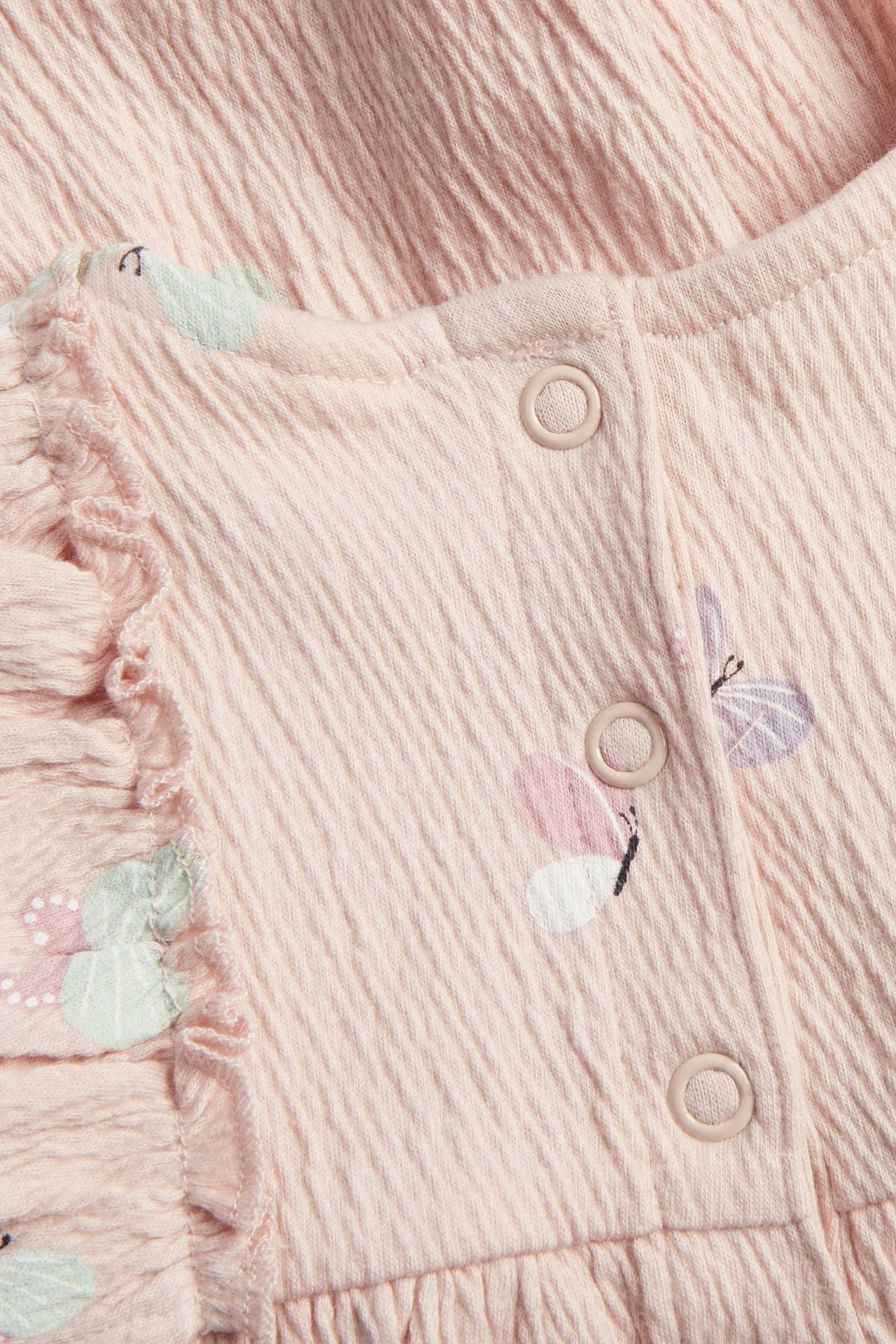 Lähikuva vauvan vaaleanpunaisesta lyhythihaisesta kreppijersey-mekosta, jossa on röyhelöitä, perhoskuvio ja painonapit.