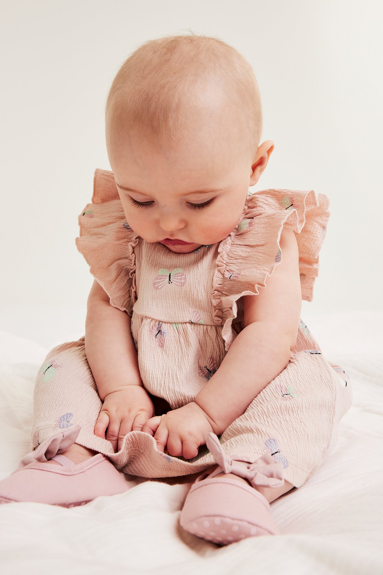 Edestä: Vauva vaaleanpunaisessa perhoskuvioidussa kreppimekossa röyhelöolkapäillä ja vaaleanpunaisilla kengillä.