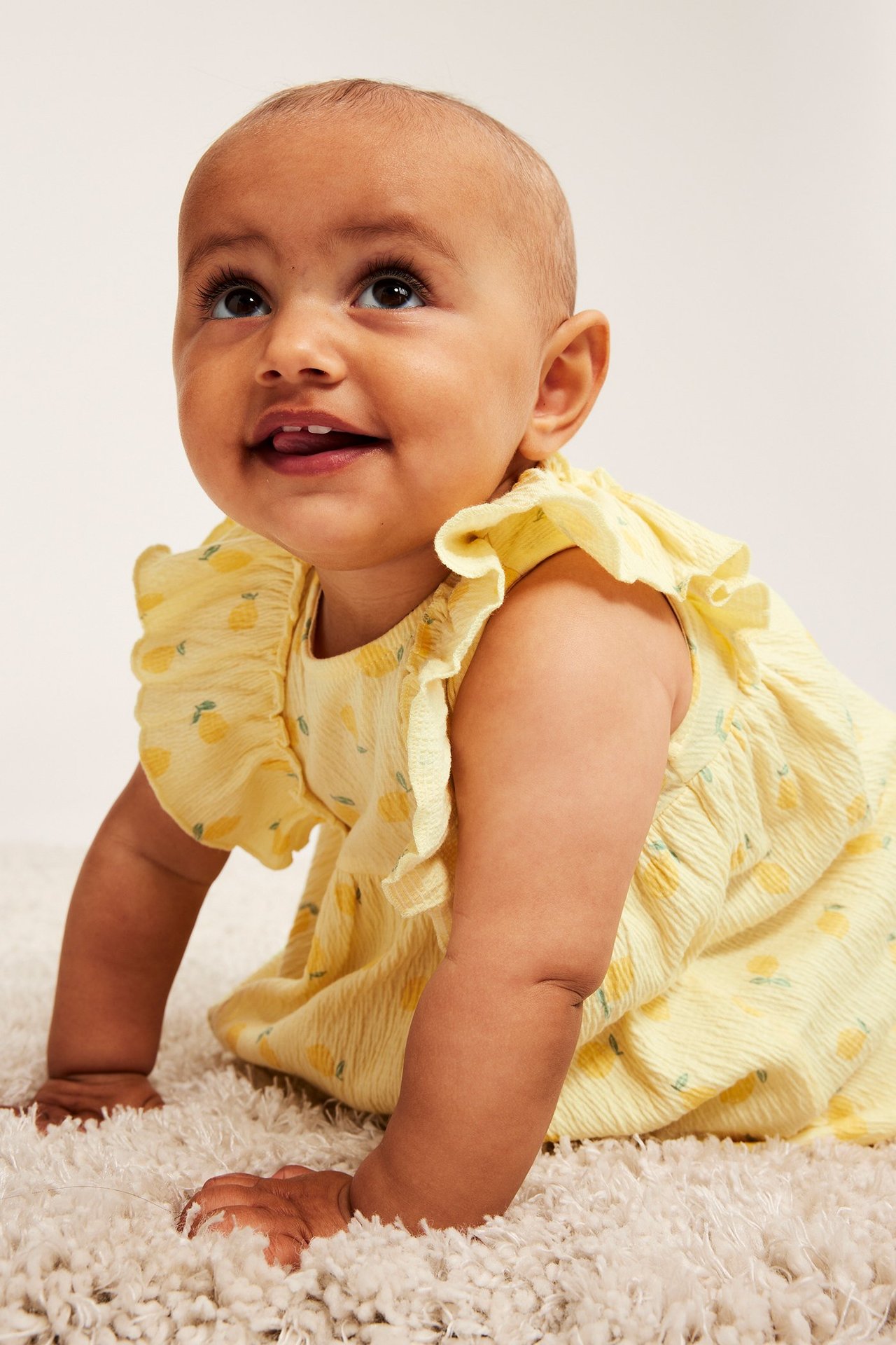 Zbliżenie na raczkujące niemowlę w jasnożółtej sukience z krepy dżersejowej w cytrynowy wzór z falbankami.