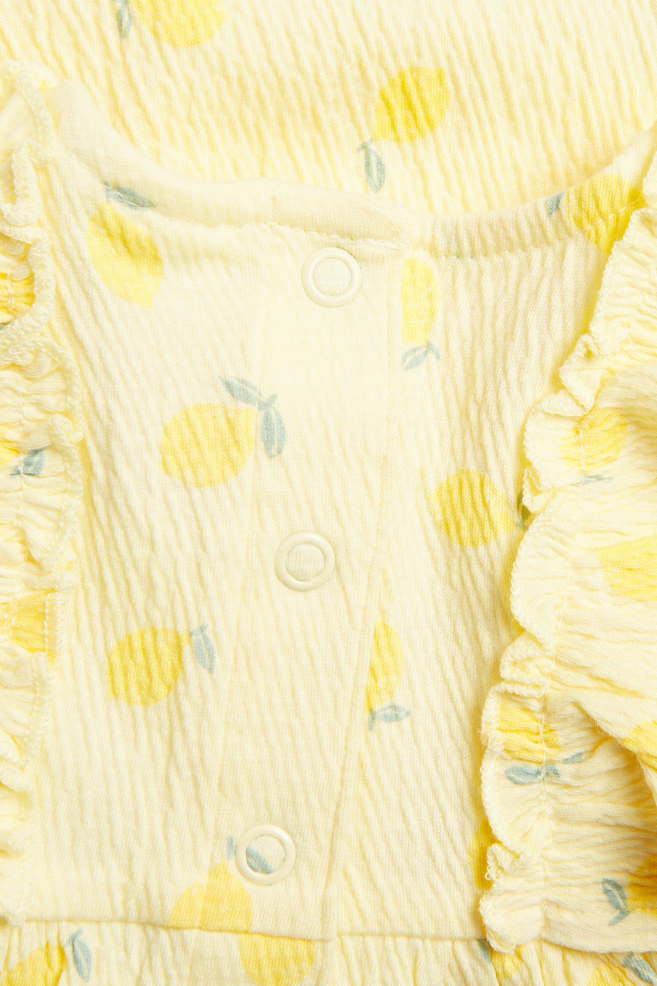 Lähikuva: vaaleankeltainen vauvan kreppijersey-mekko sitruunaprintillä, röyhelöillä ja painonapeilla edessä.