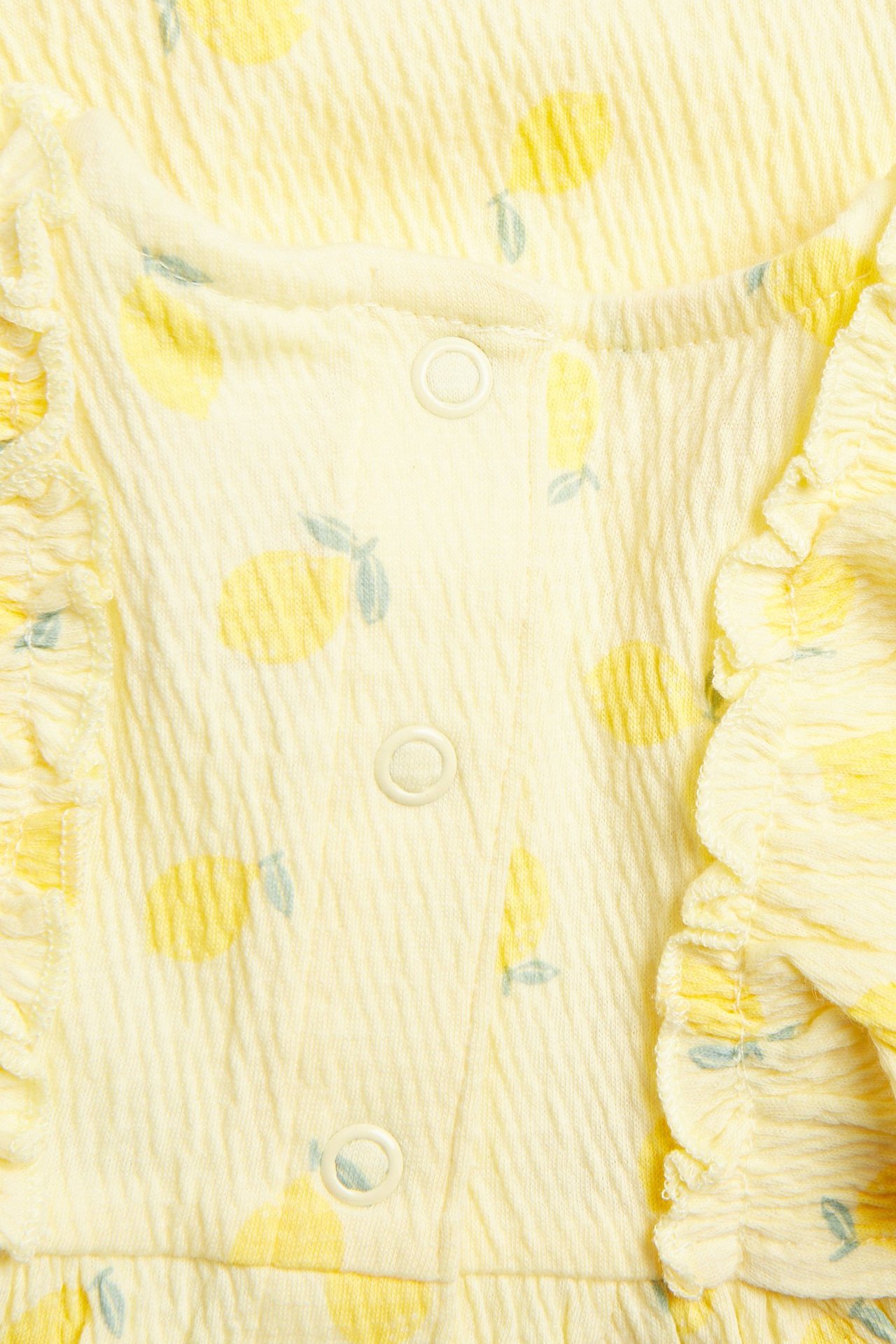 Lähikuva: vaaleankeltainen vauvan kreppijersey-mekko sitruunaprintillä, röyhelöillä ja painonapeilla edessä.
