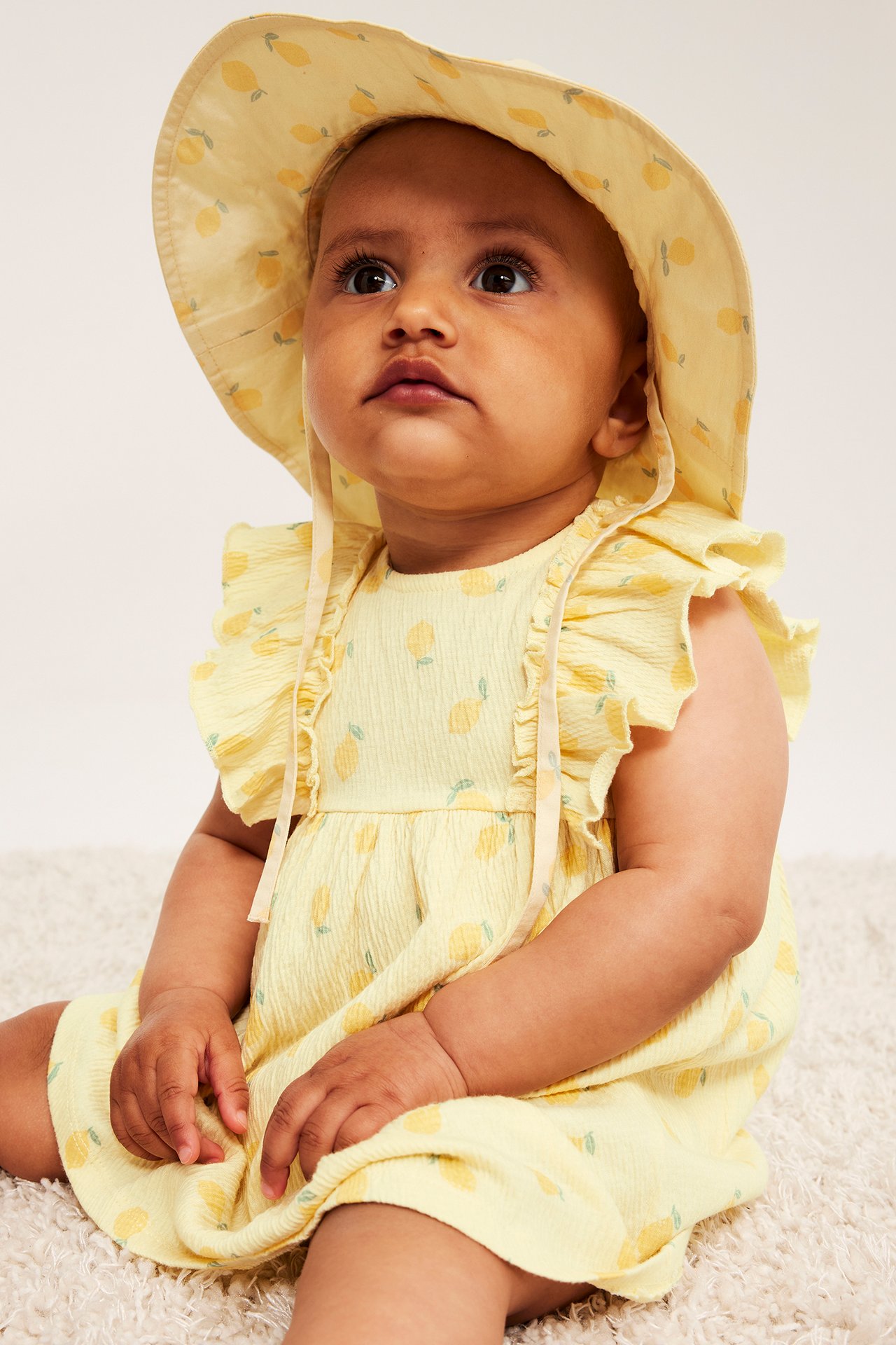 Zbliżenie: Dziecko nosi jasnożółtą krepową sukienkę w cytryny z falbankami i pasujący kapelusz.