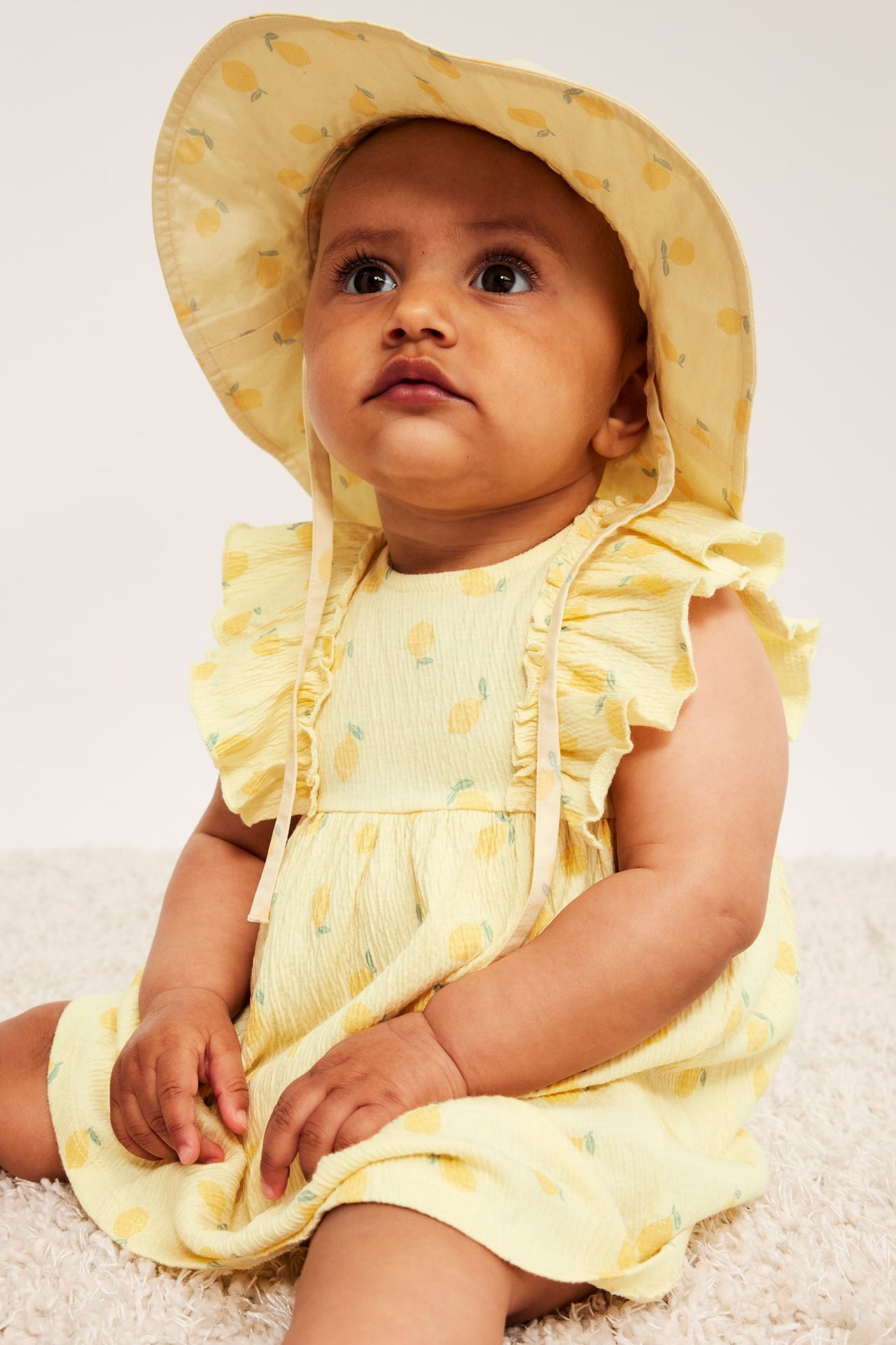 Zbliżenie: Dziecko nosi jasnożółtą krepową sukienkę w cytryny z falbankami i pasujący kapelusz.
