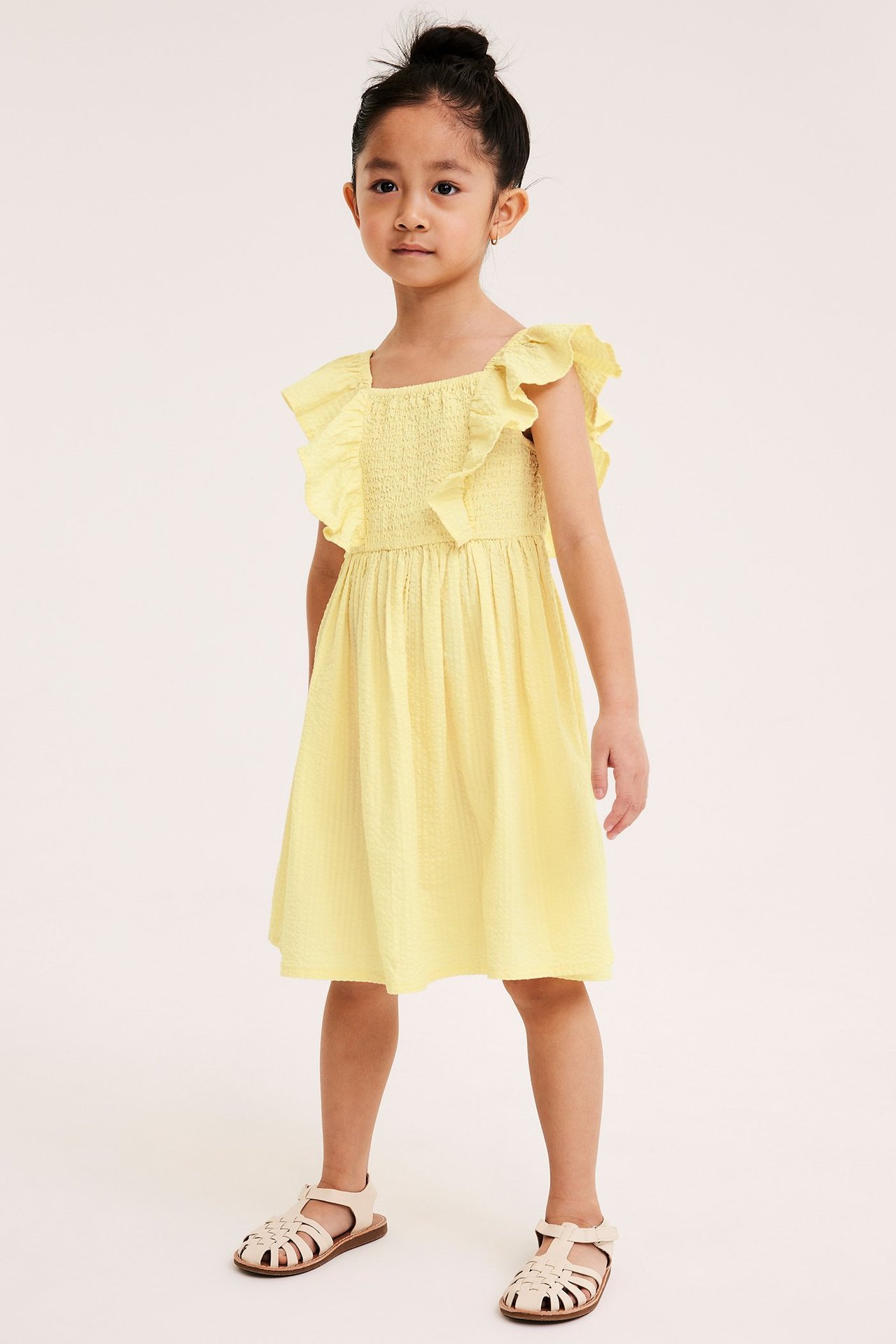 Widok z przodu: dziecko w jasnożółtej bawełnianej sukience z marszczeniami, falbankami i beżowych sandałach.