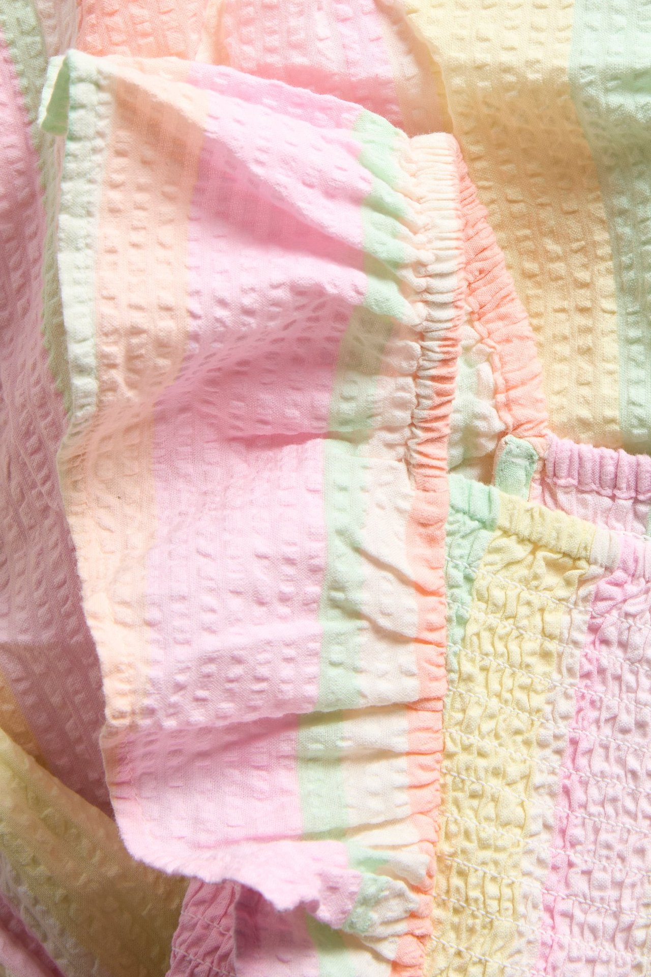 Zbliżenie: Dziecięca sukienka z teksturowanej bawełny w pastelowe paski, z falbanką i marszczeniami.